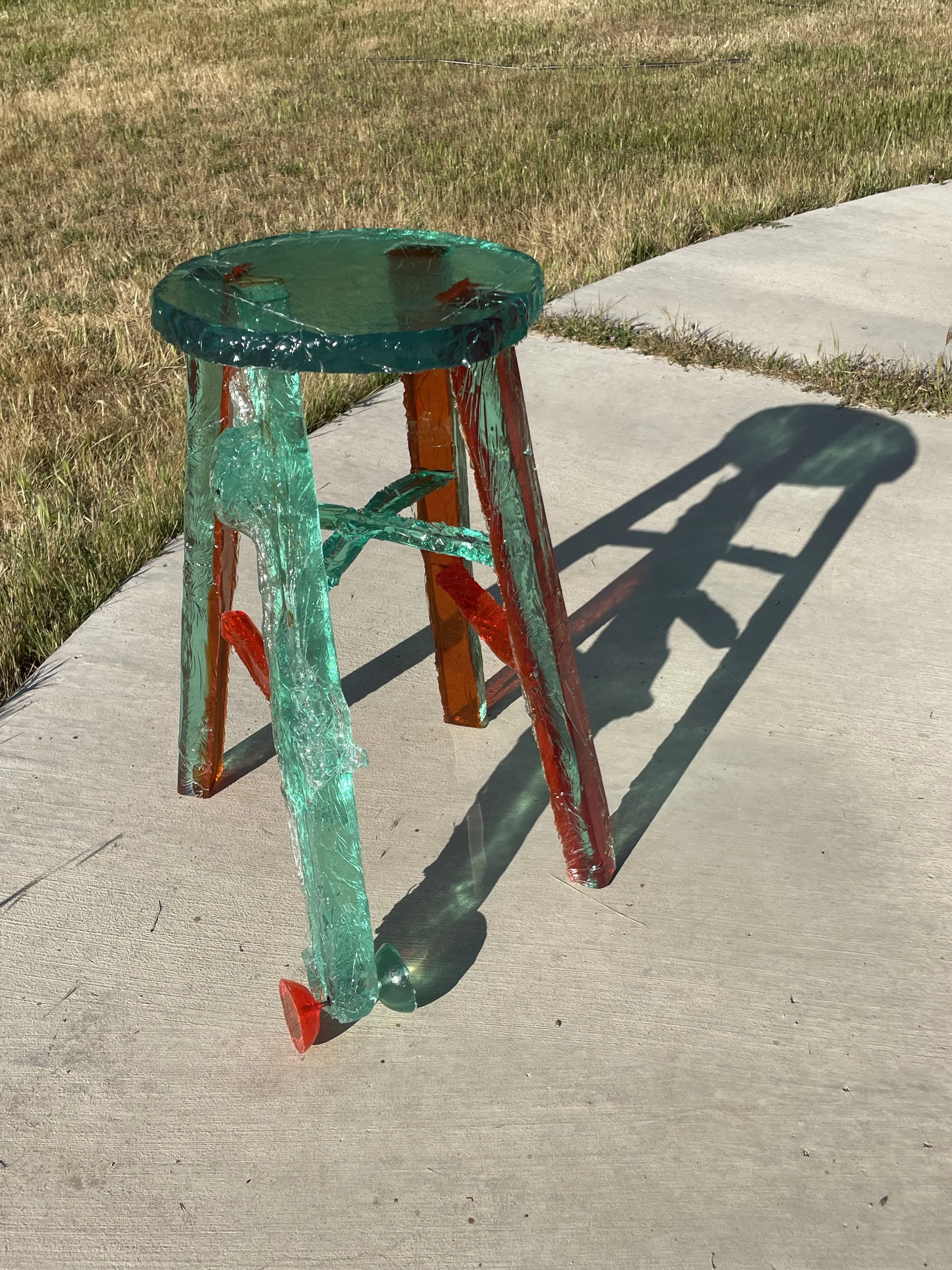 A glass sculpture of a stool on a concrete sidewalk, casting a shadow on the ground.