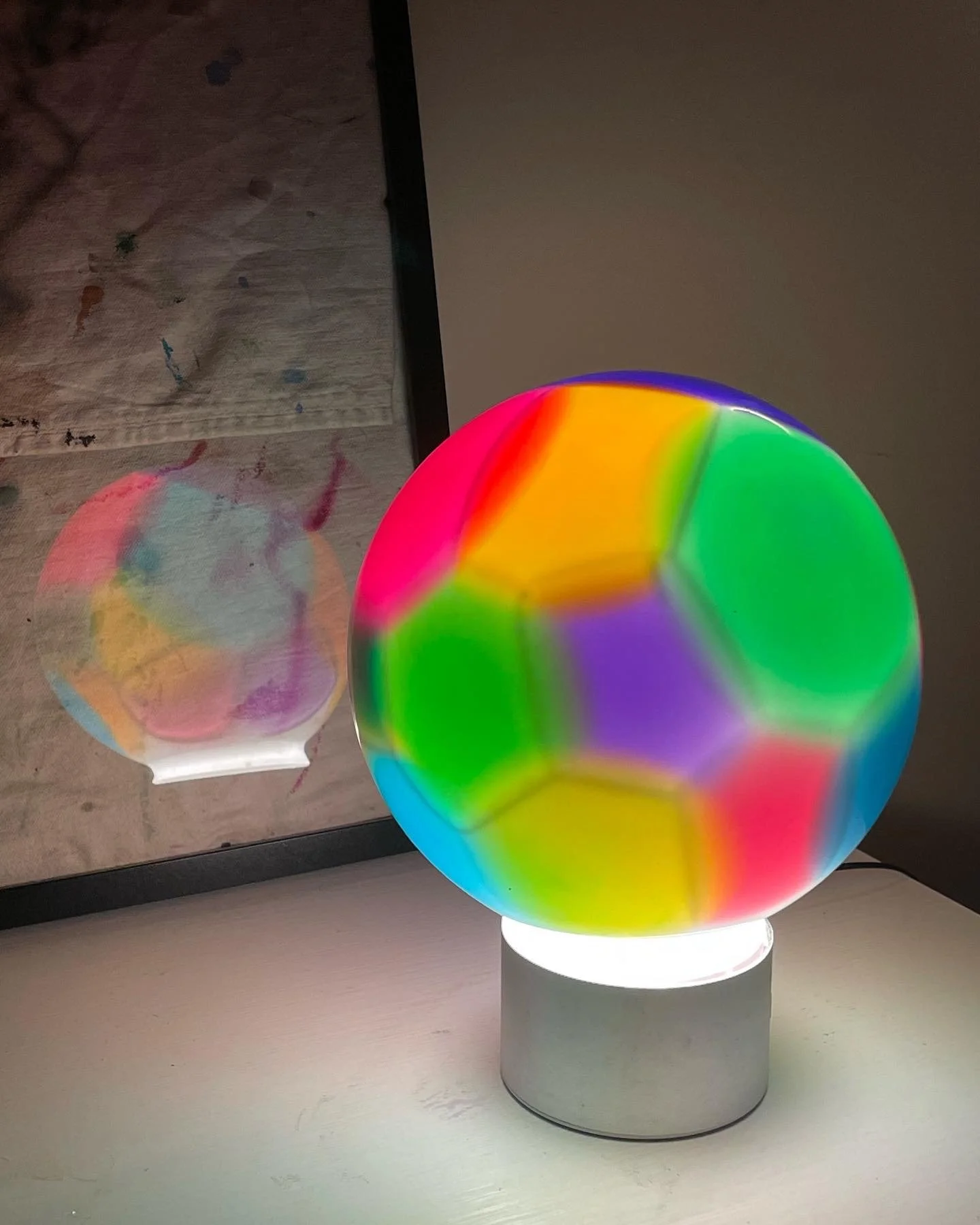 Colorful illuminated spherical resin lamp with a reflection of the same lamp's on a picture frame behind it.
