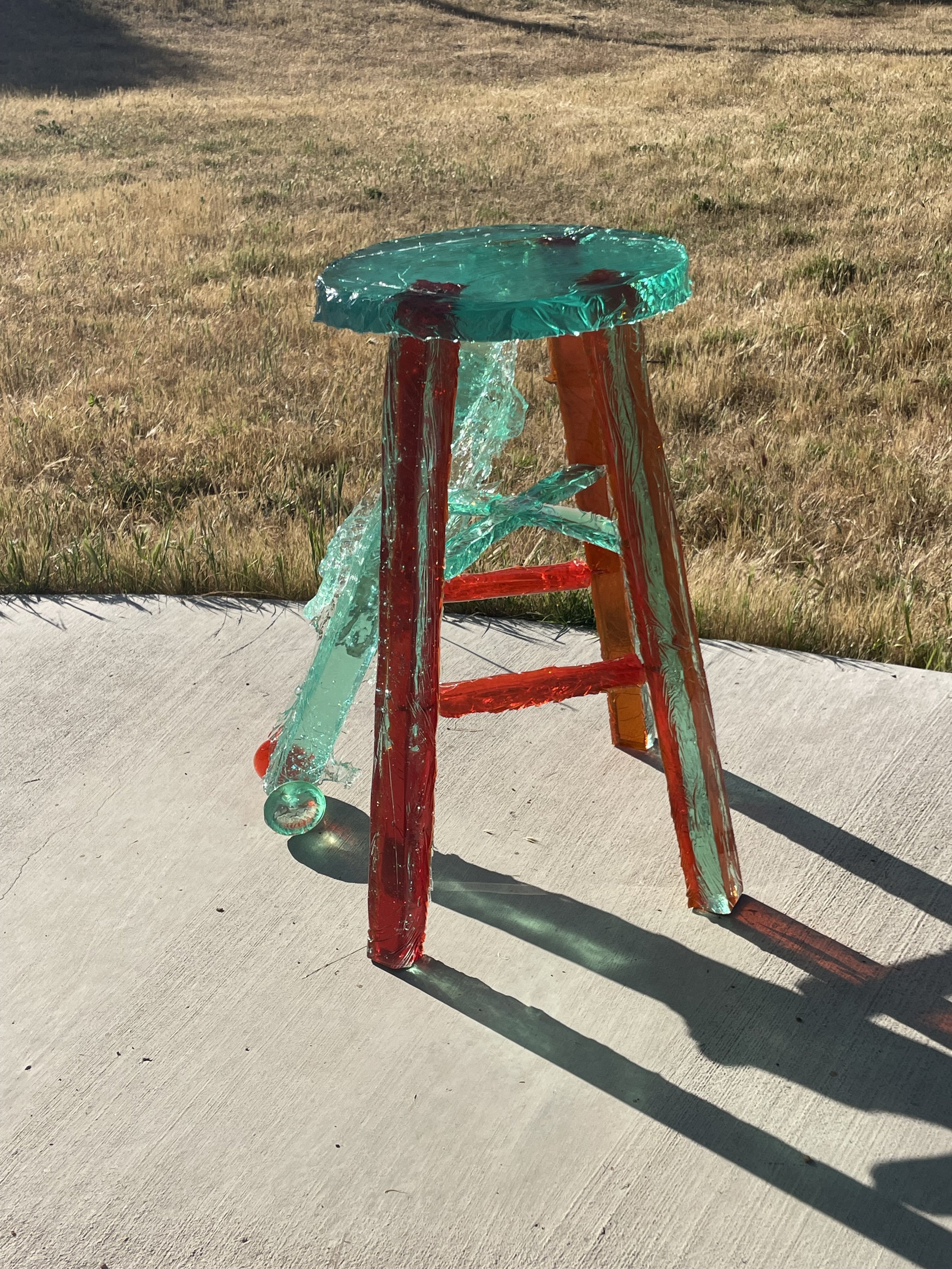 A broken glass stool with red legs and a round seat, shattered and lying on a sidewalk outside on a sunny day.