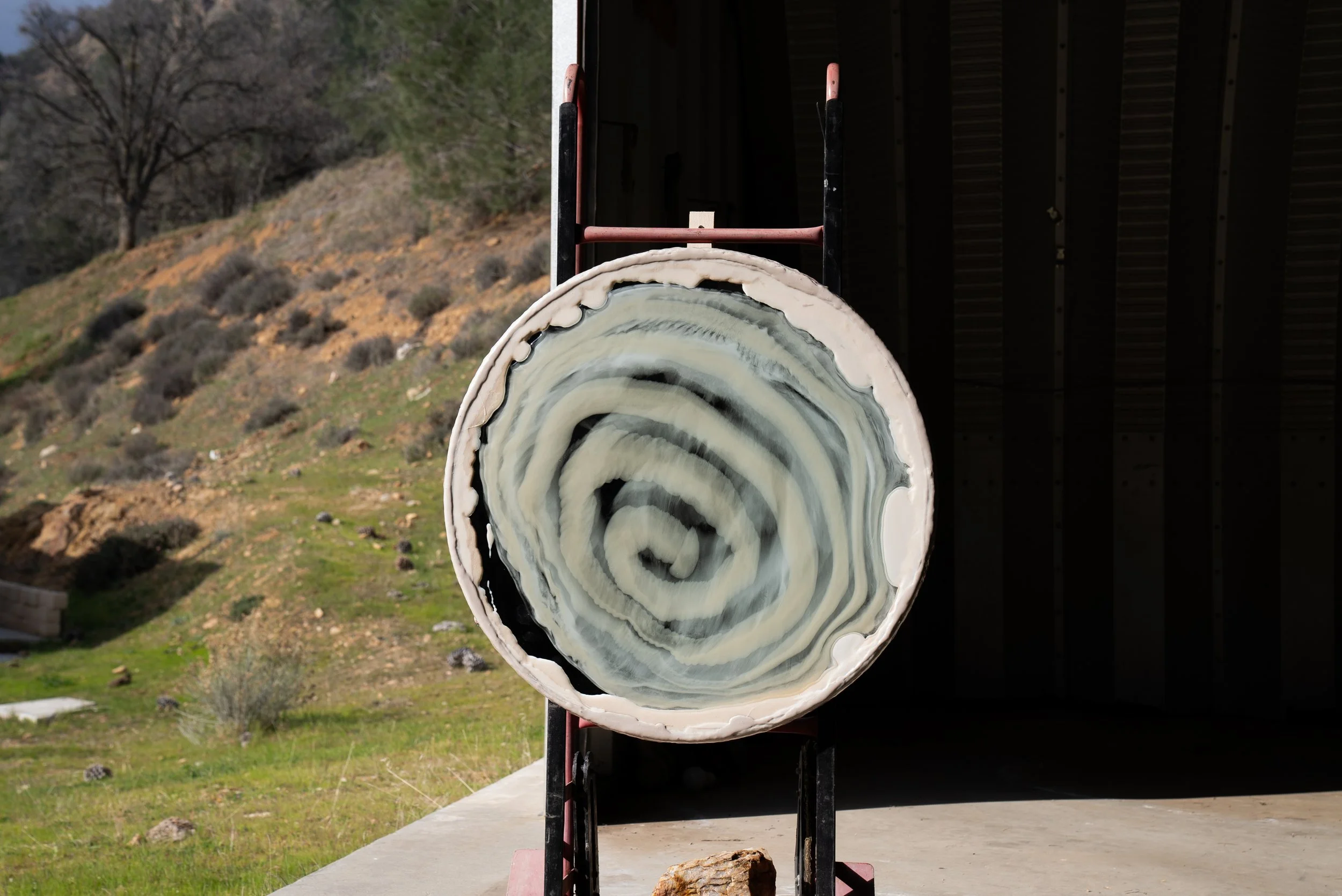 A large, circular resin mirror with white and clear swirling patterns, mounted on a pink and black stand outside, with mountains and trees in the background.
