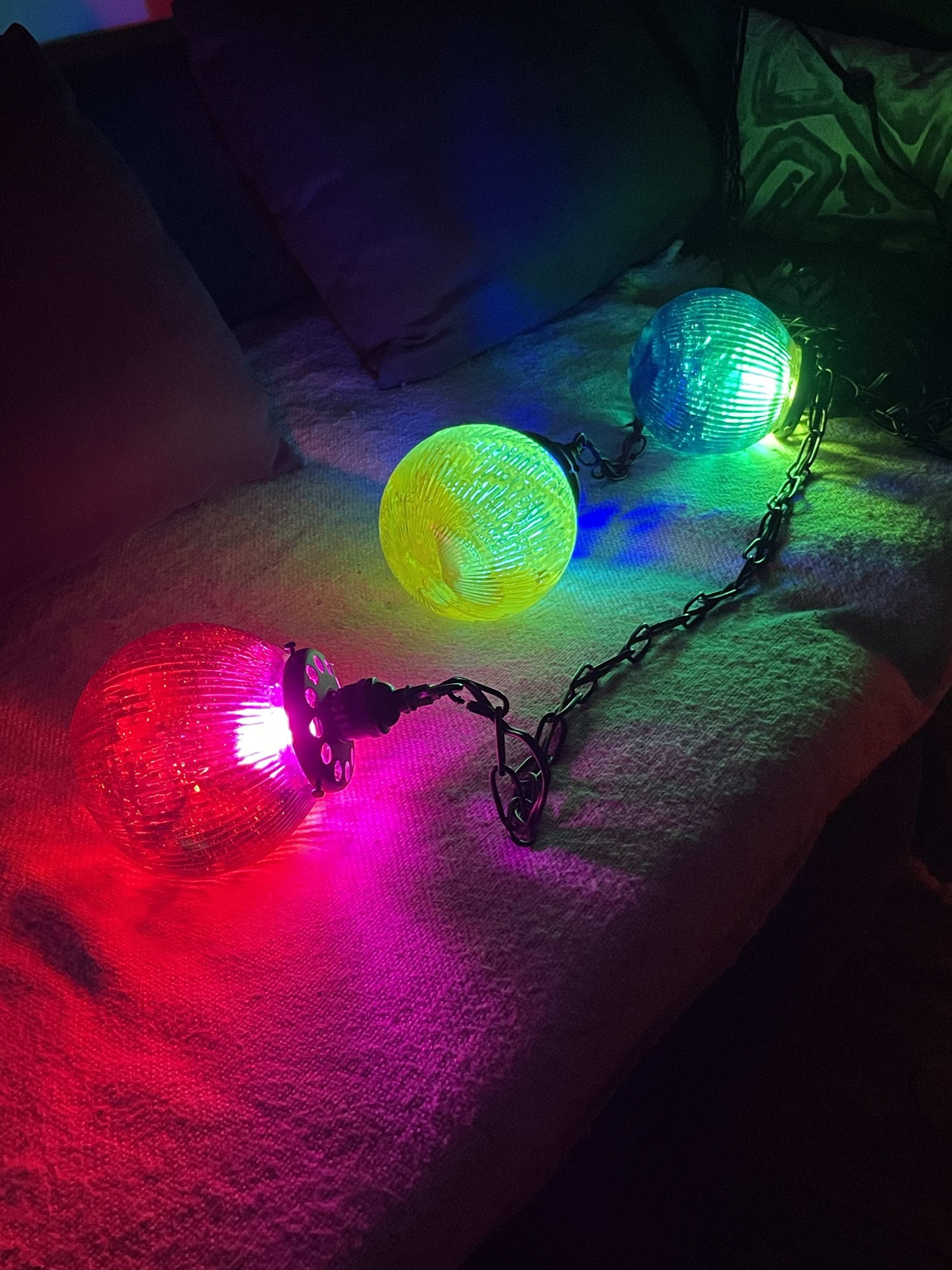 Colorful resin lights with three spherical covers in red, yellow, and blue are arranged on a plush couch with purple cushions, casting colorful reflections.