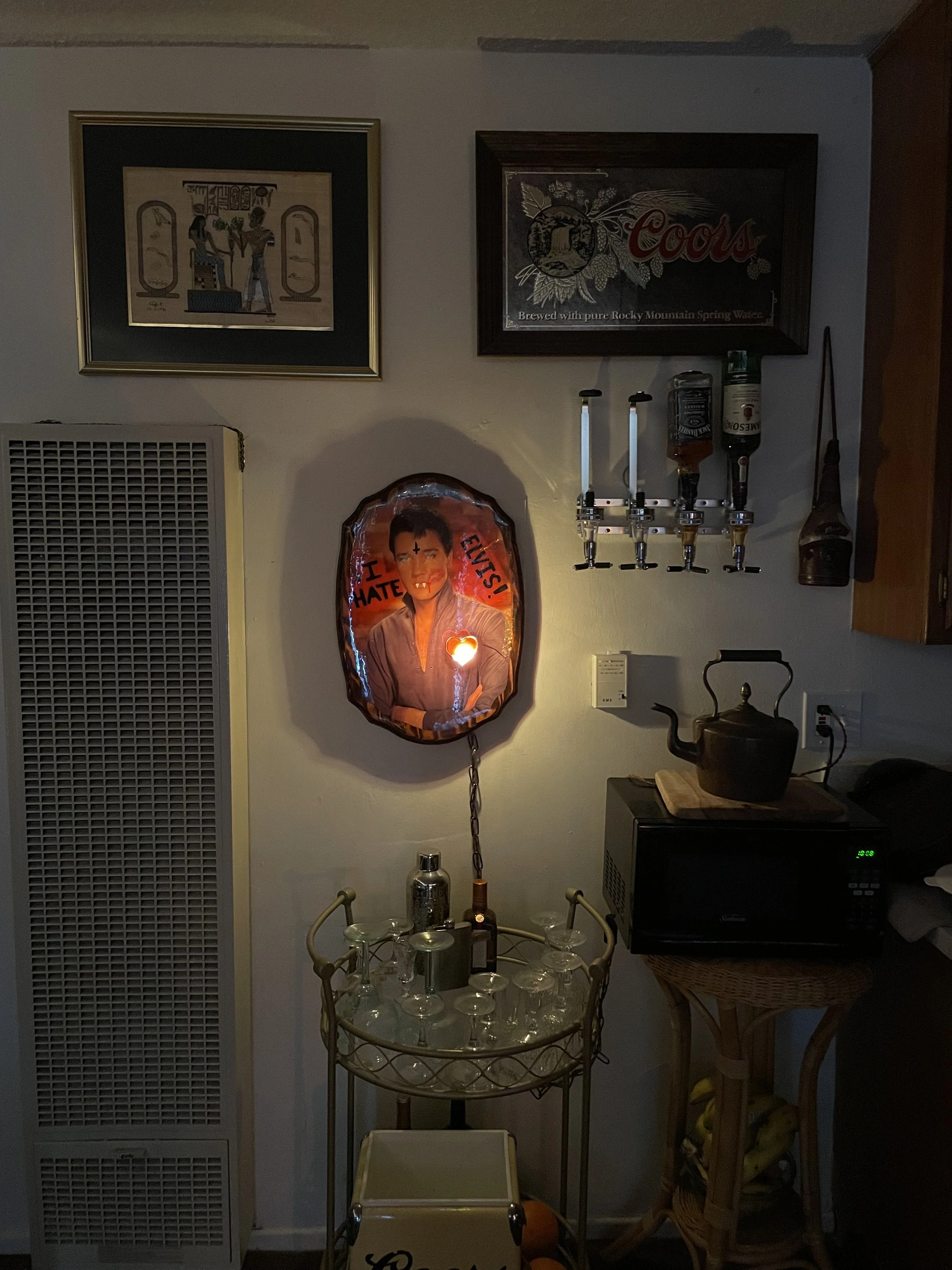 A dimly lit kitchen corner with vintage decor, including a Elvis Presley portrait with I HATE ELVIS!! text and a heart shaped resin light, framed artwork, a bar with alcohol bottles, a microwave, a teapot, a small table, and bananas.