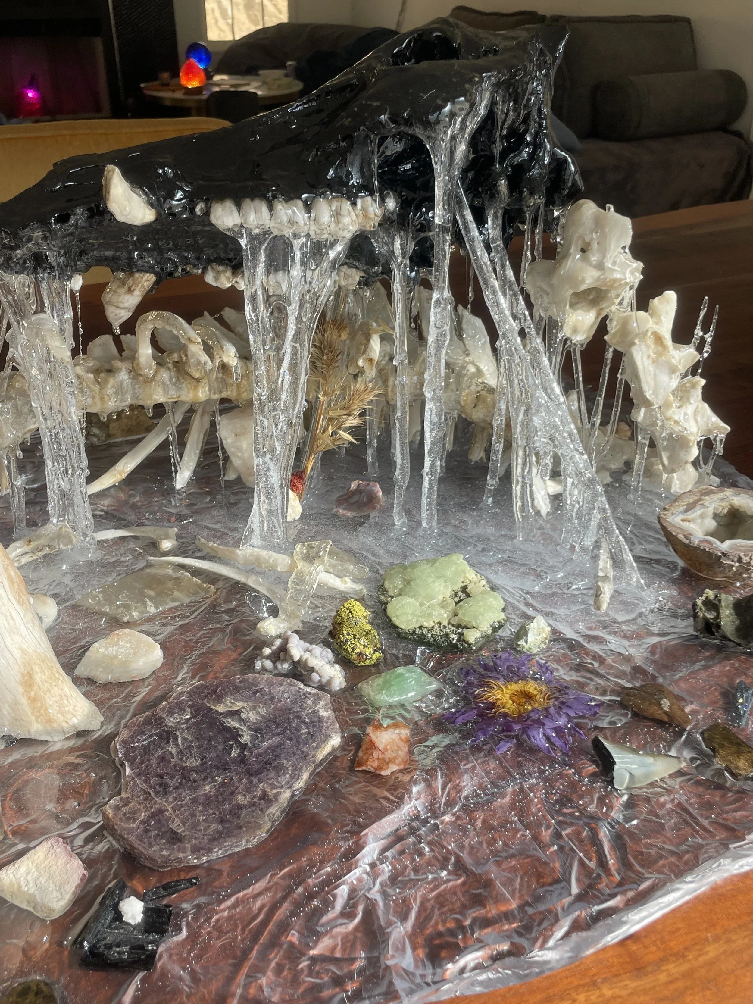A skull sculpture with resin icicles hanging from it, surrounded by various rocks, minerals, and decorative objects set in resin on a wooden table.