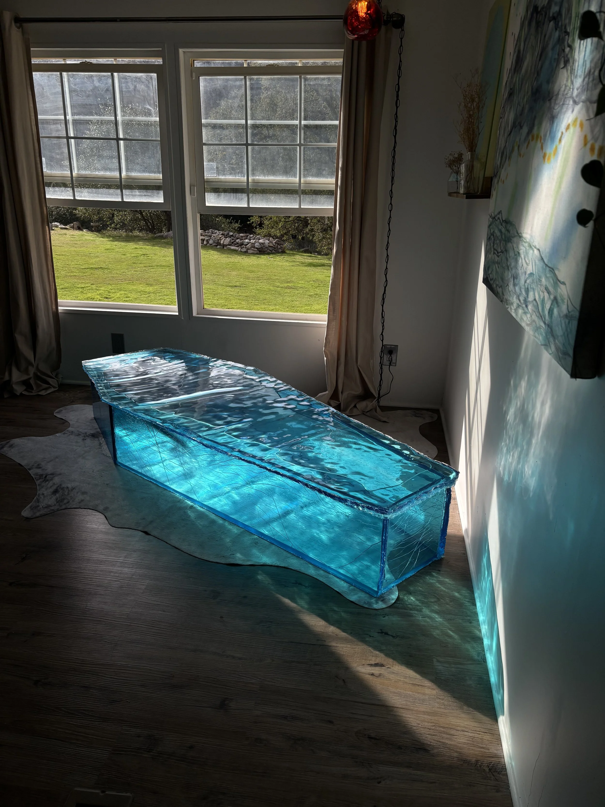 A clear blue resin coffin coffee table, placed on a cowhide rug in front of large windows with beige curtains, letting in natural light. Outside is a grassy backyard with trees and rocks.