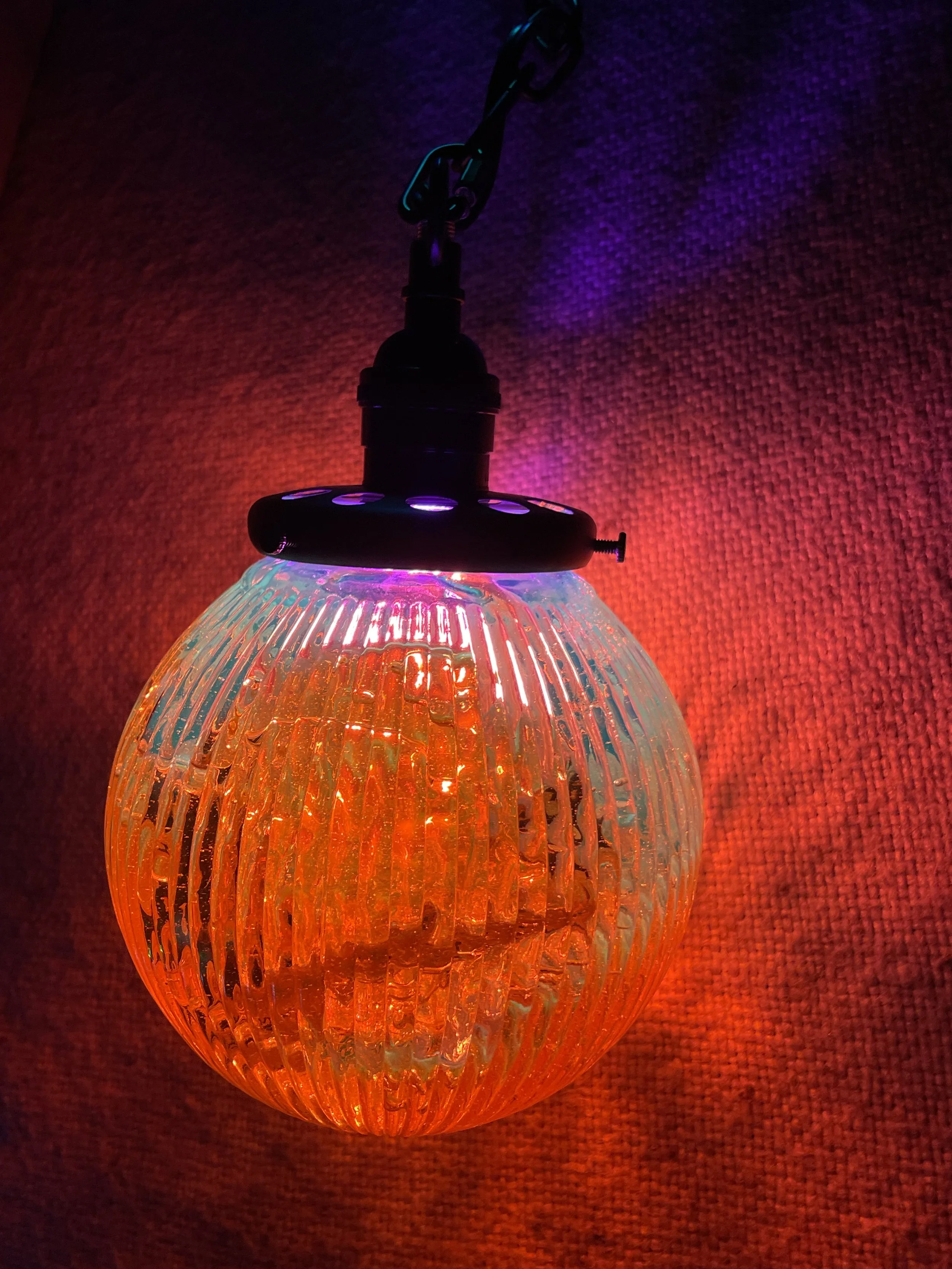 Colorful hanging resin lamp with ribbed design emitting red, orange, and purple, light against a textured wall.
