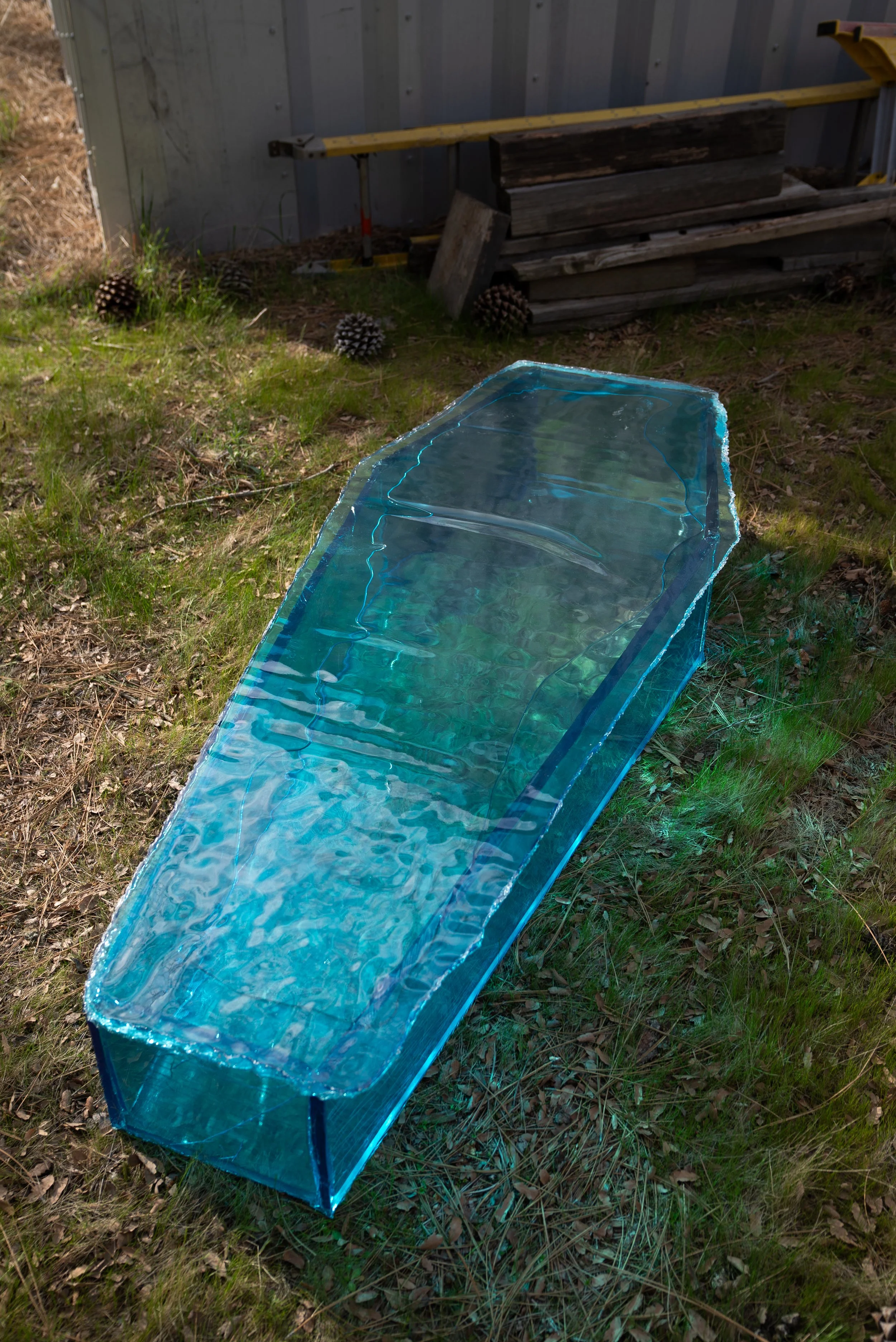 A clear blue resin coffin resting on grass outdoors near a weathered building and stack of wood.