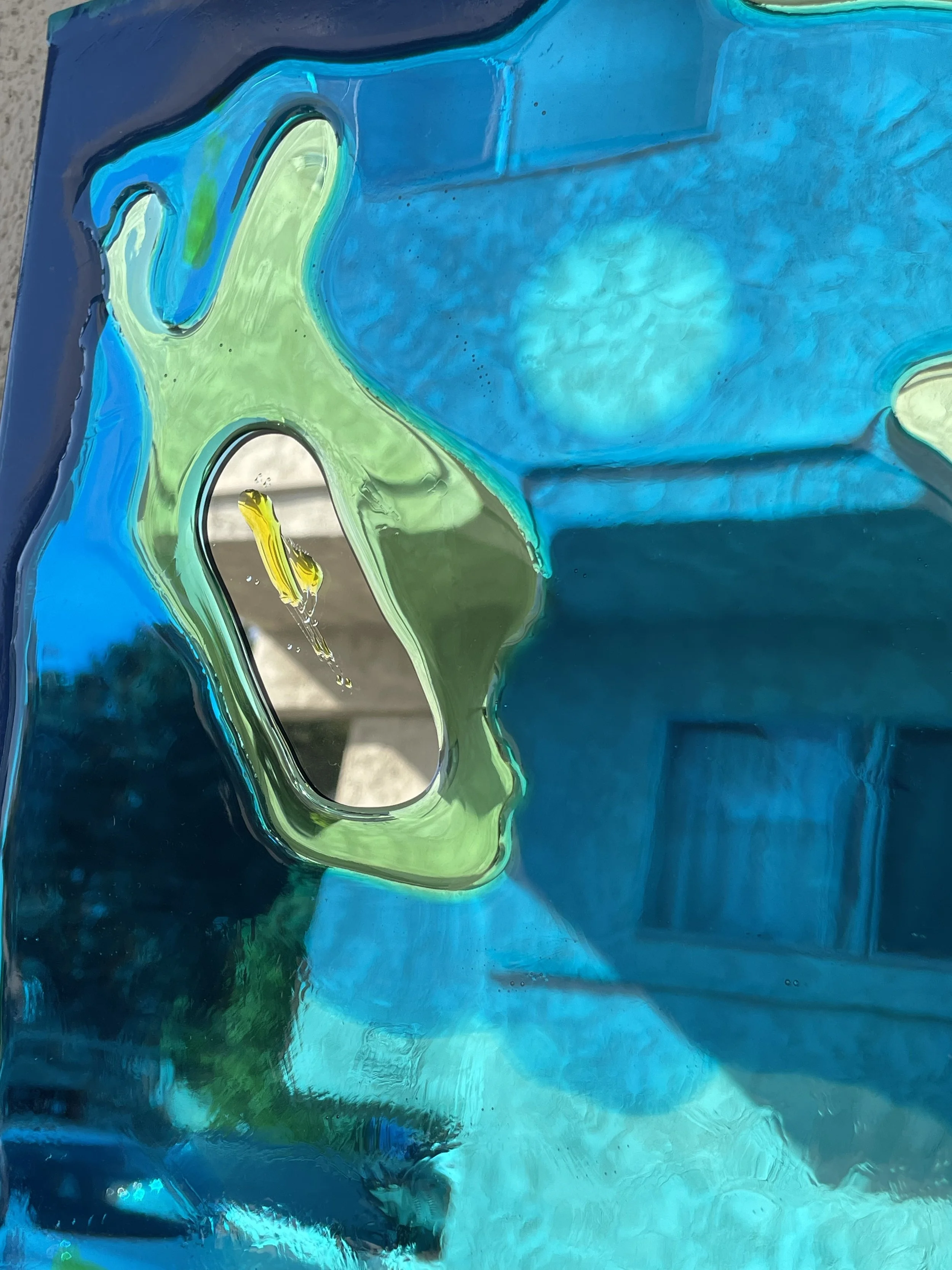 Close-up of a blue and green abstract resin artwork with a reflective surface, showing distortions and reflections of the surrounding environment.