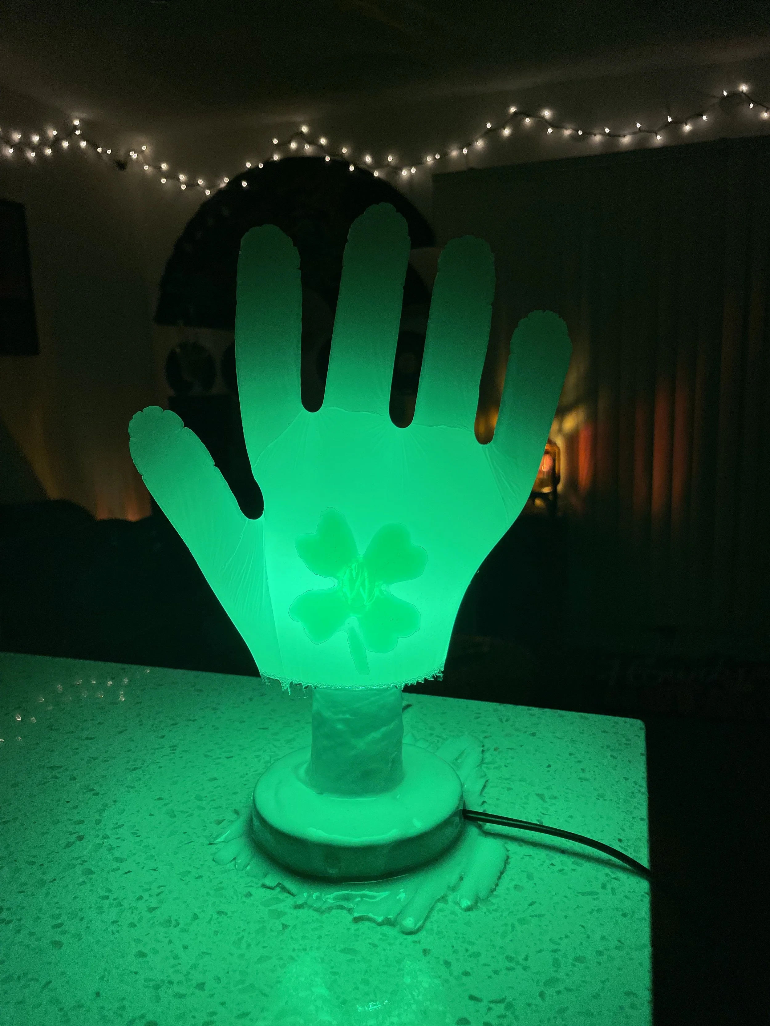 A white resin decorative lamp in the shape of a hand with a shamrock in the palm, emitting green light, on a textured table in a dimly lit room with string lights on the ceiling.