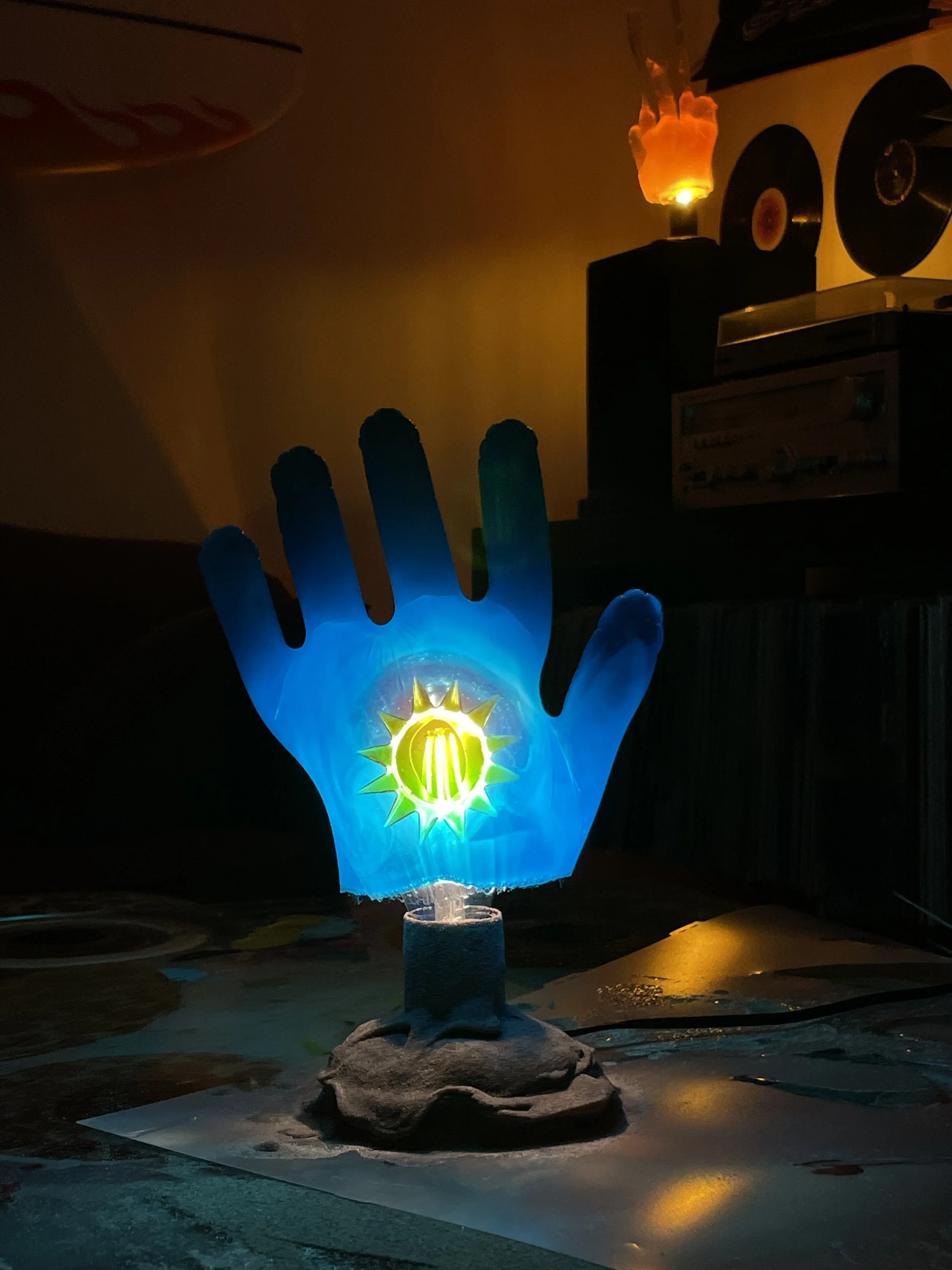 A glowing resin light in the shape of a hand with a sun design on the palm, placed on a sand base, with a dark background.