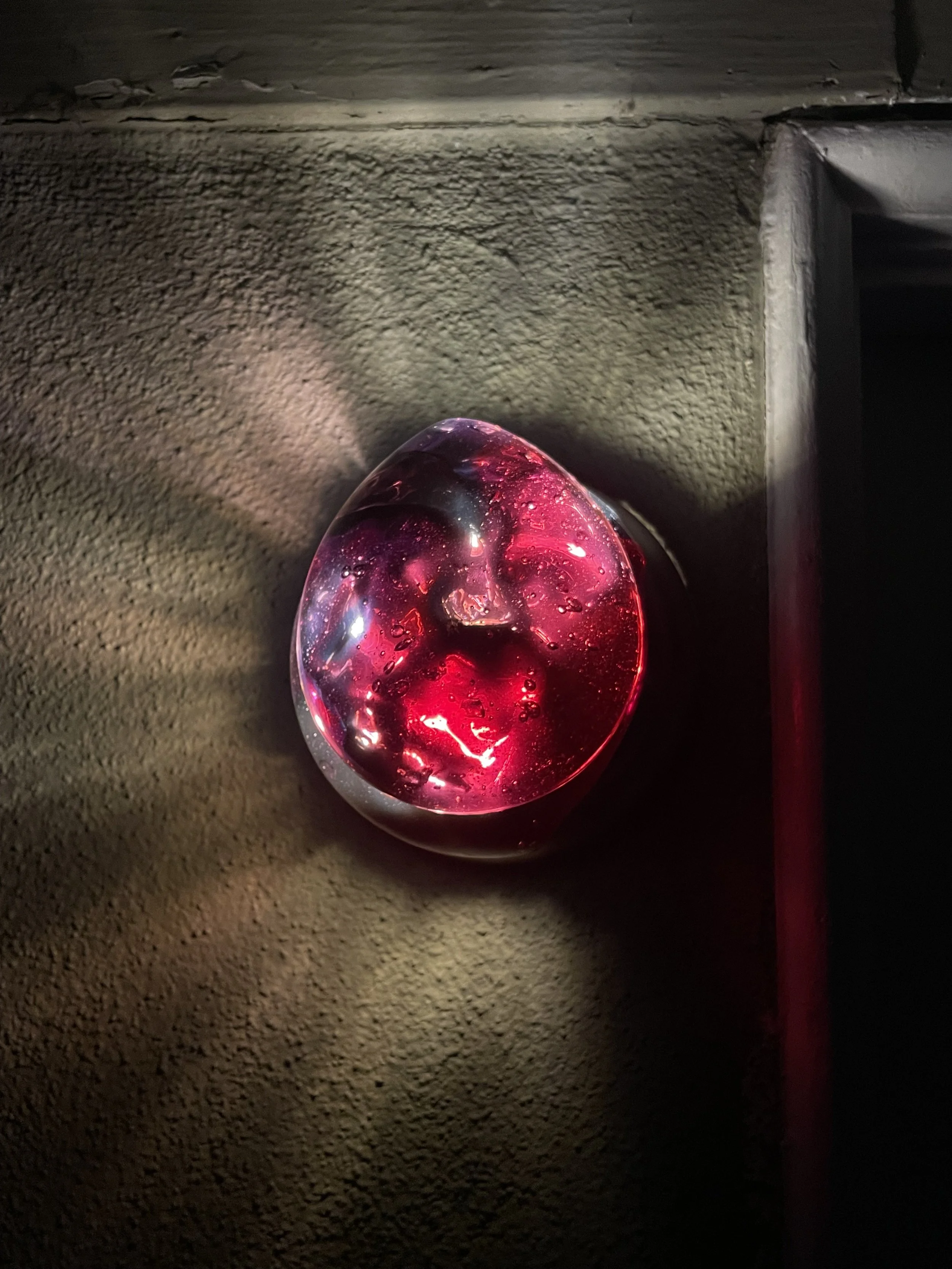 A resin light sculpture with a red hue, resembling a face or mask, illuminated and mounted on a textured wall.
