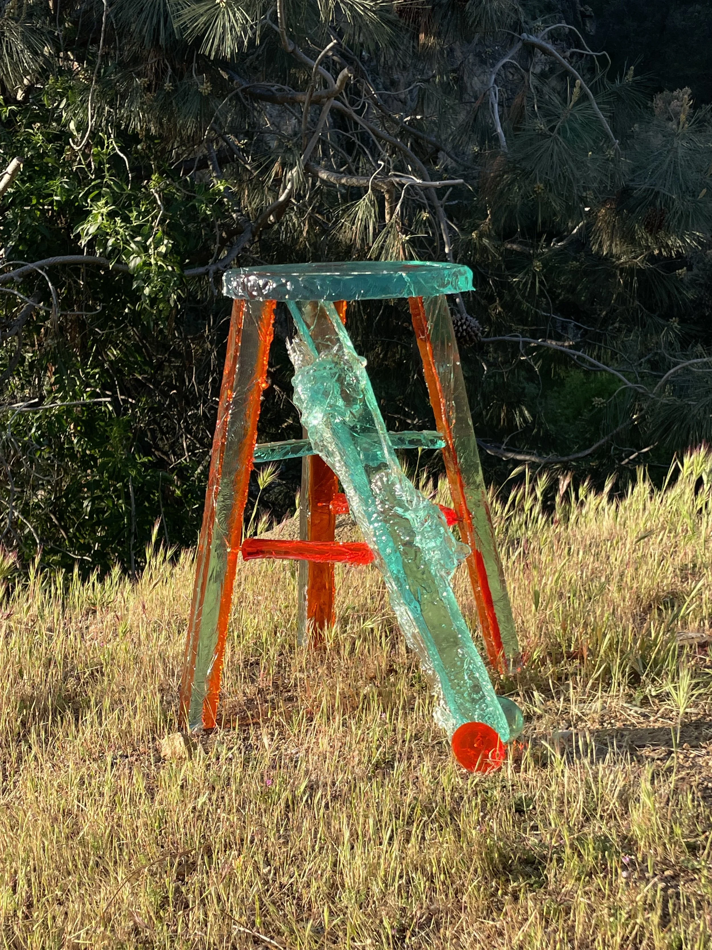 An outdoor scene with a resin stool colored in green, orange, and red. The resin appears to be melted or broken, hanging on a grassy area with trees in the background.