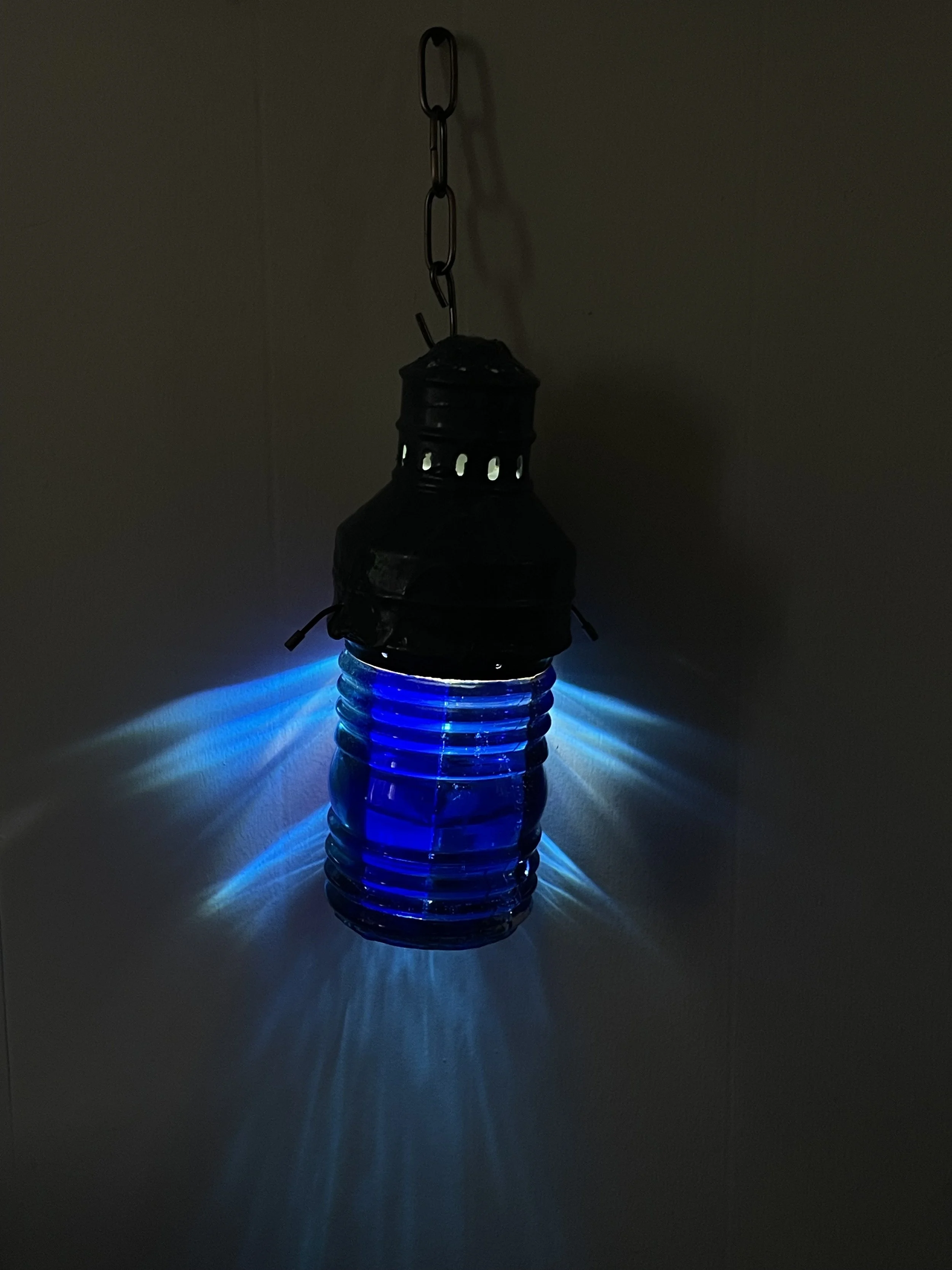 An upcycled lantern made from a glass jar filled with blue resin, with a metal top, hanging from a chain on a wall, illuminated to project blue light and pattern shadows on the wall.