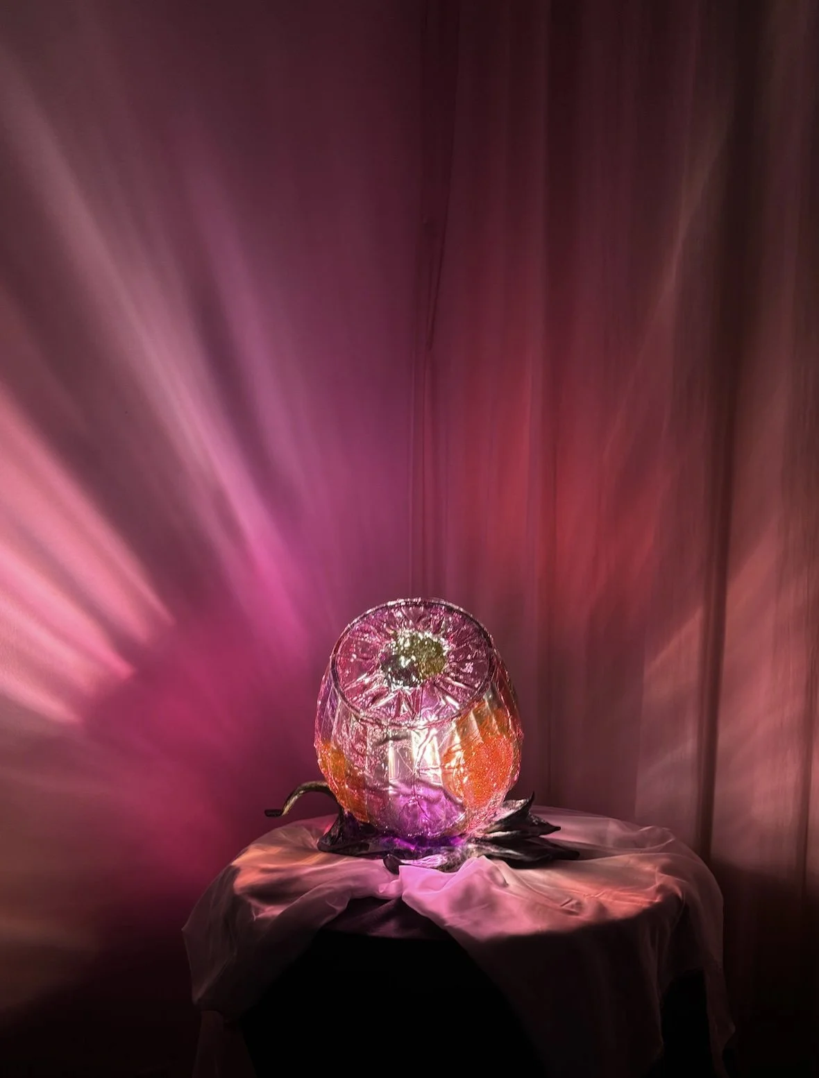Ice like resin rose sculpture illuminated with pink and purple lights, placed on a table with a cloth, against a pink backdrop.