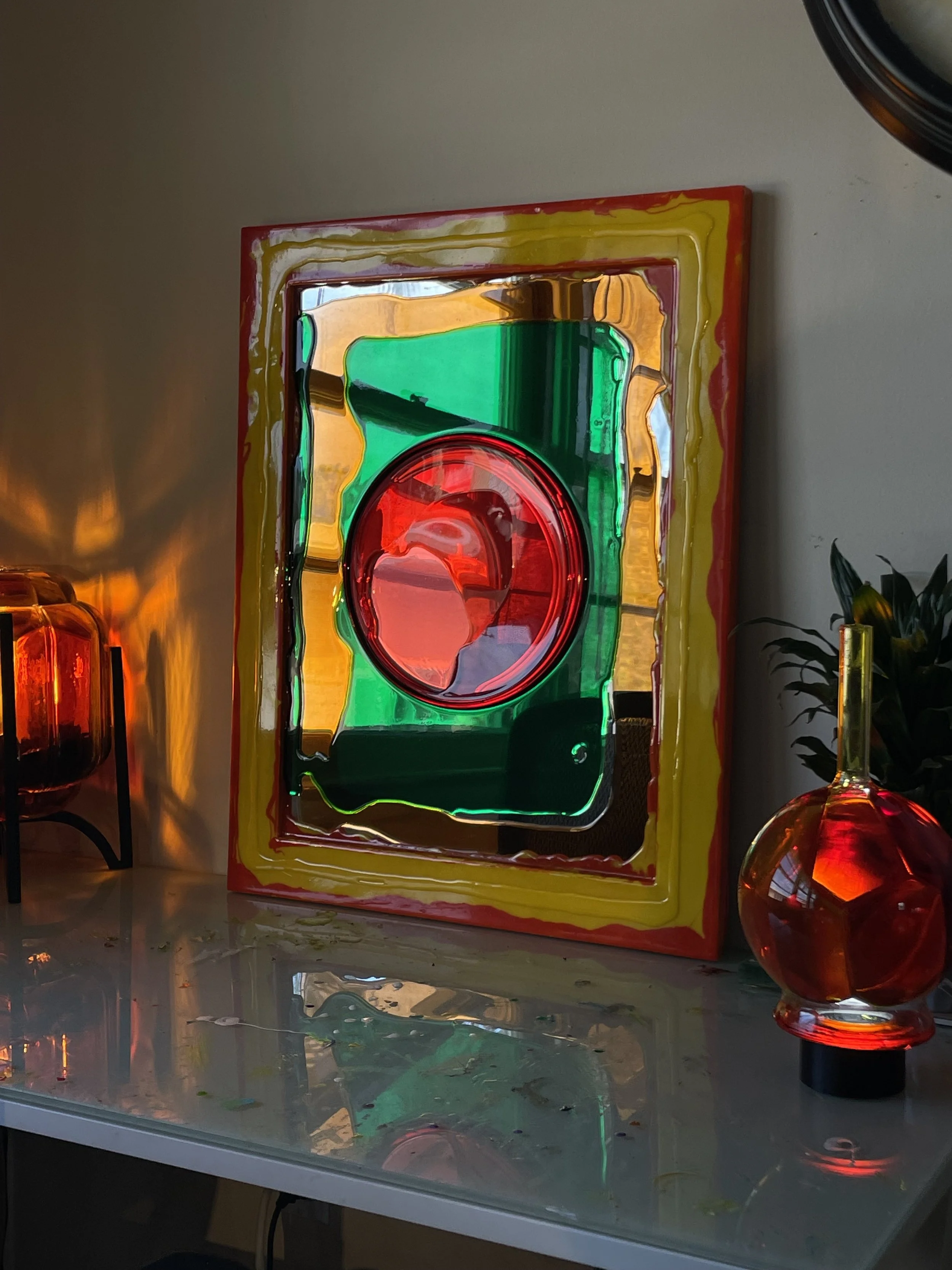 Colorful abstract resin mirror with red, green, yellow, and orange segments, placed on a reflective surface, with a decorative red resin and glass object and a plant to the side.