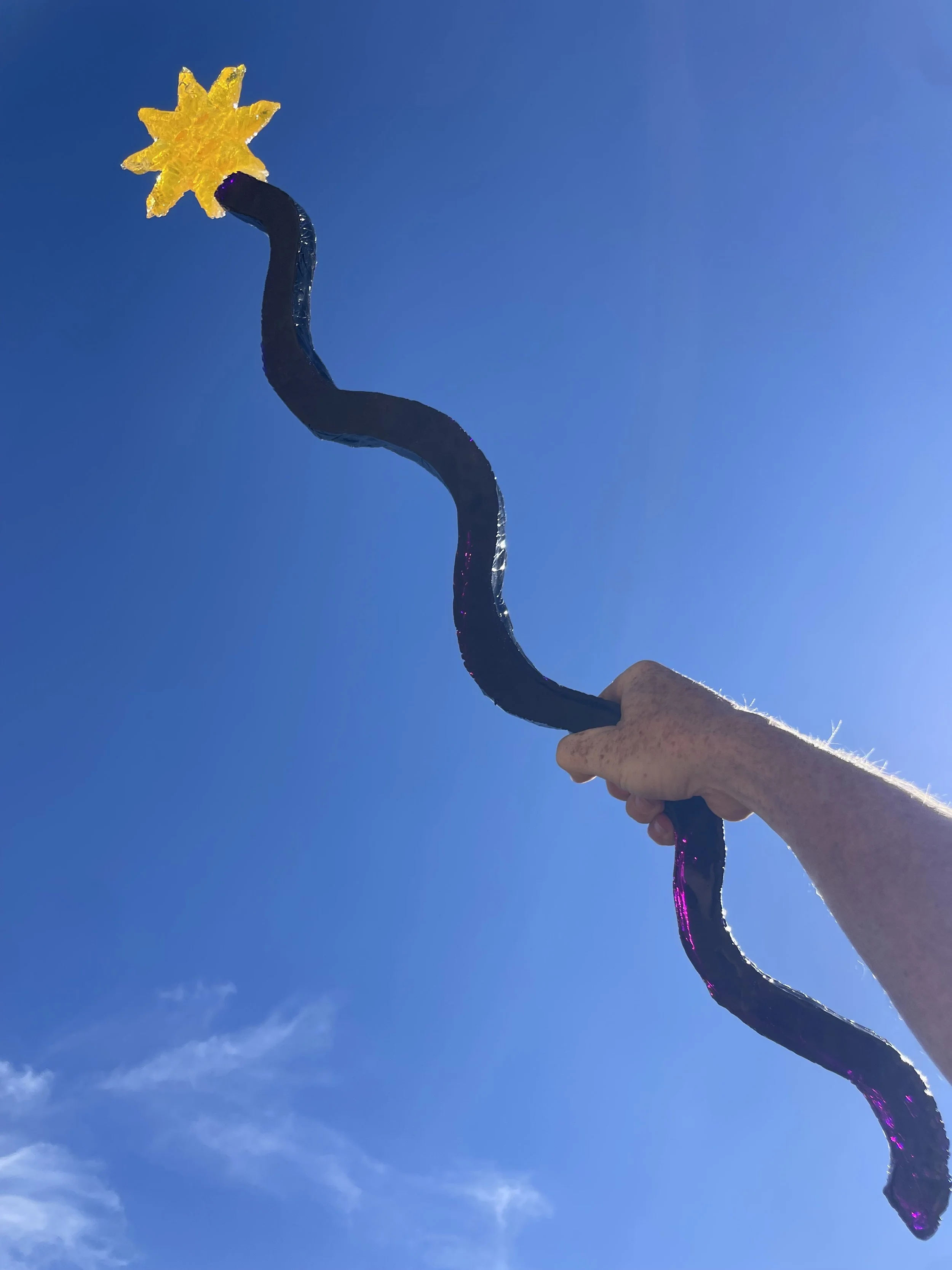 A hand holding a dark purple, twisted resin magic wand with a yellow sun-shaped top against a bright blue sky.