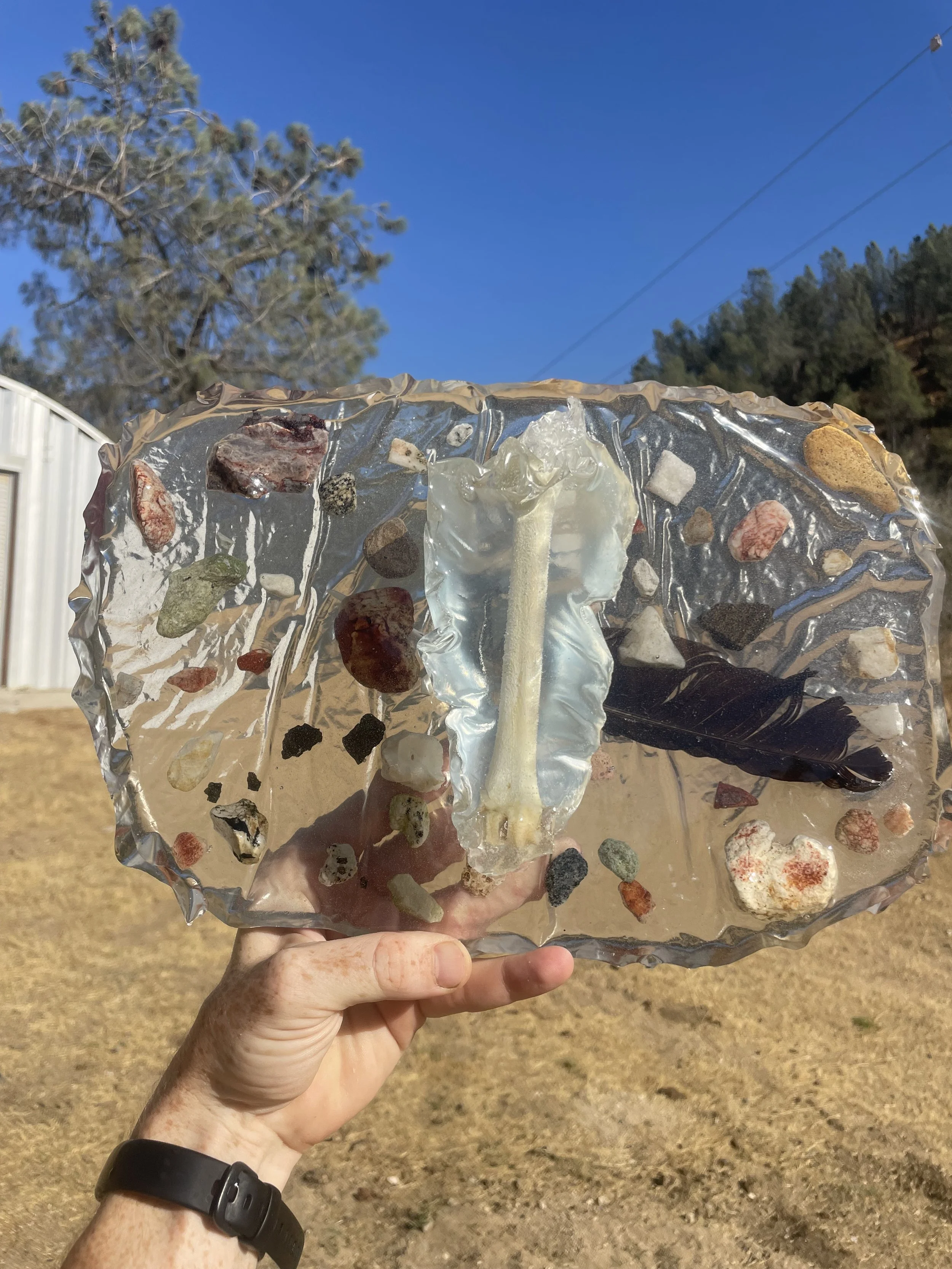A person holding a piece of clear resin with small rocks, a feather, and a bone embedded inside, outdoors under a blue sky with trees and a white building in the background.