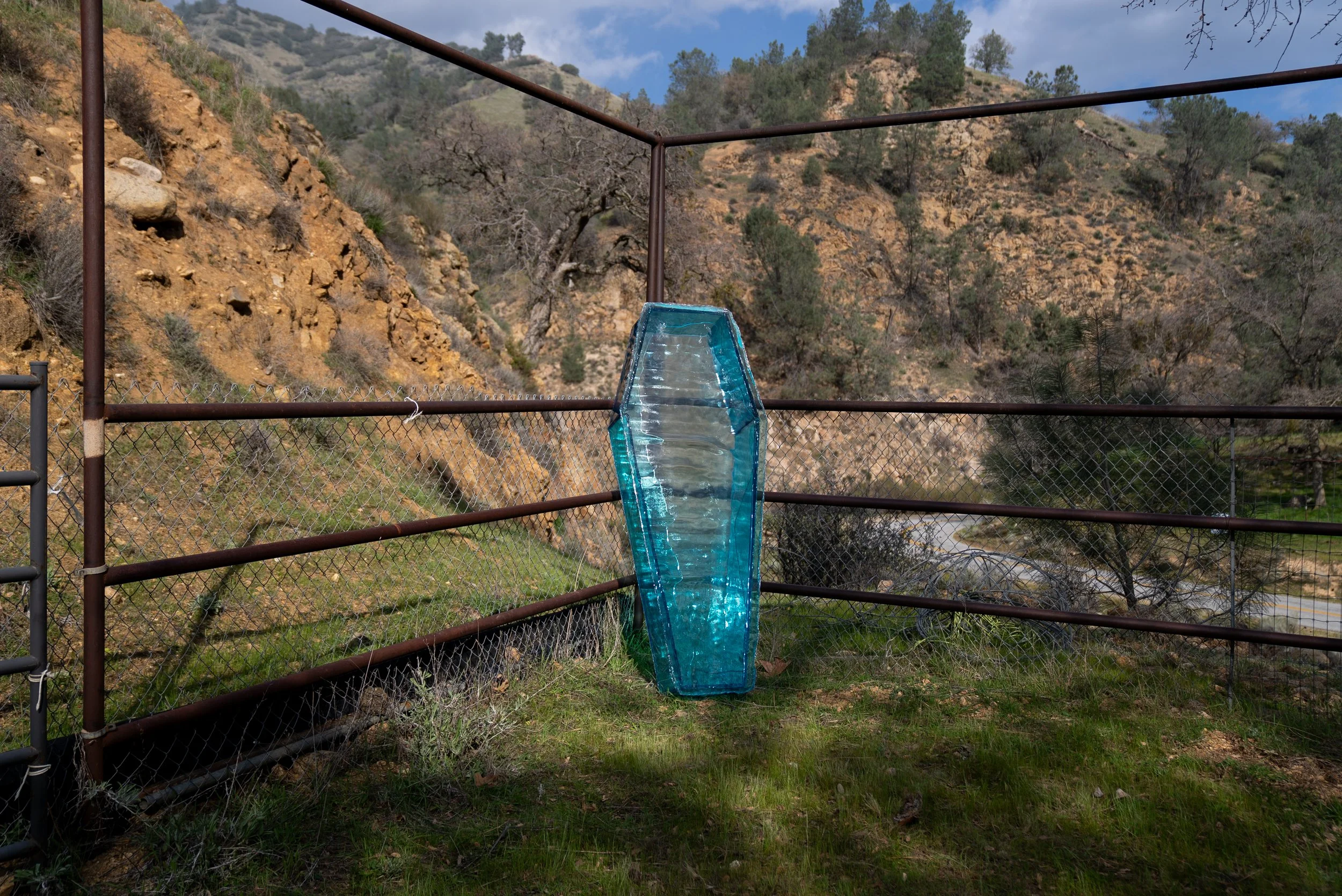 A blue, coffin-shaped resin sculpture leaning against a chain-link fence, with a mountainous hillside in the background.