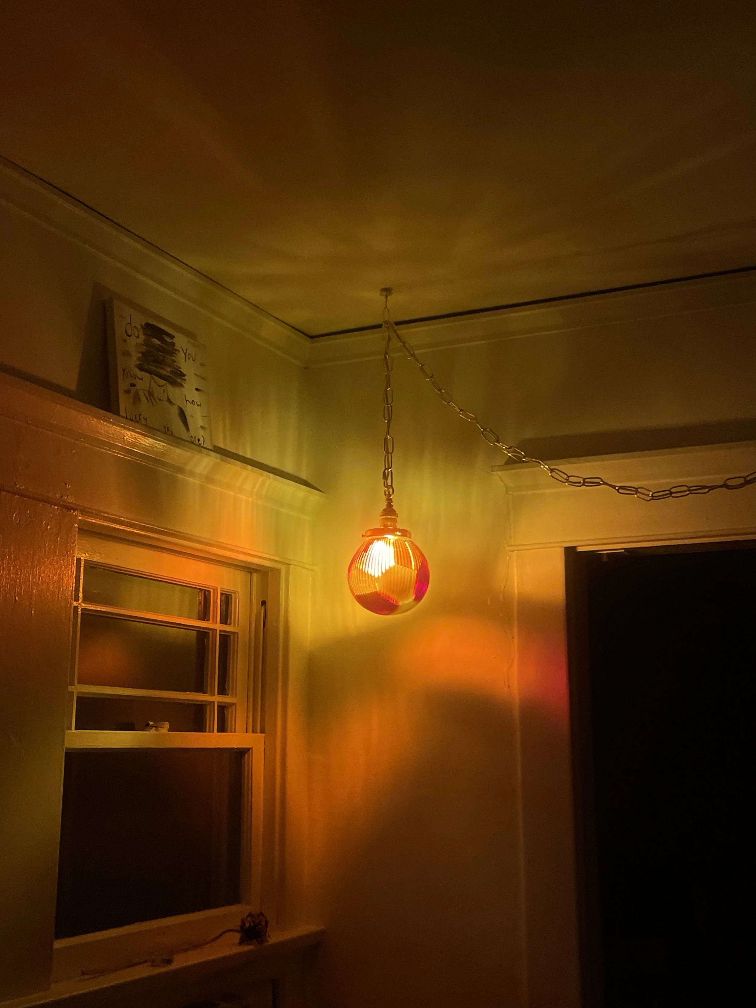 A ceiling light fixture with a reddish-orange resin lamp hanging from a chain in a dimly lit room, casting warm reflections on the wall.