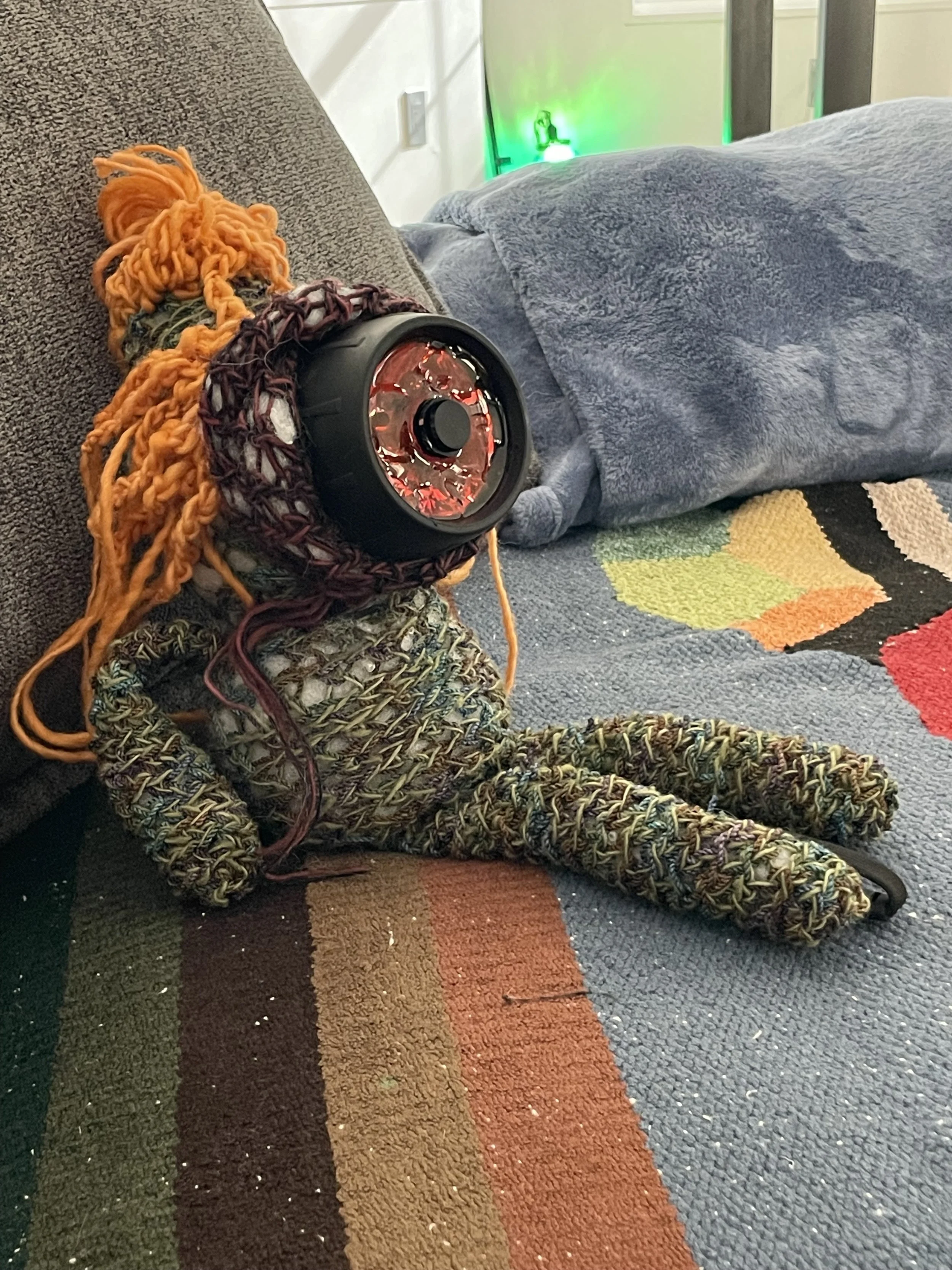 A colorful crocheted puppet with a resin-customized diving lamp in its face, sitting on a couch with a patterned blanket. There is a green glowing light on the wall in the background.