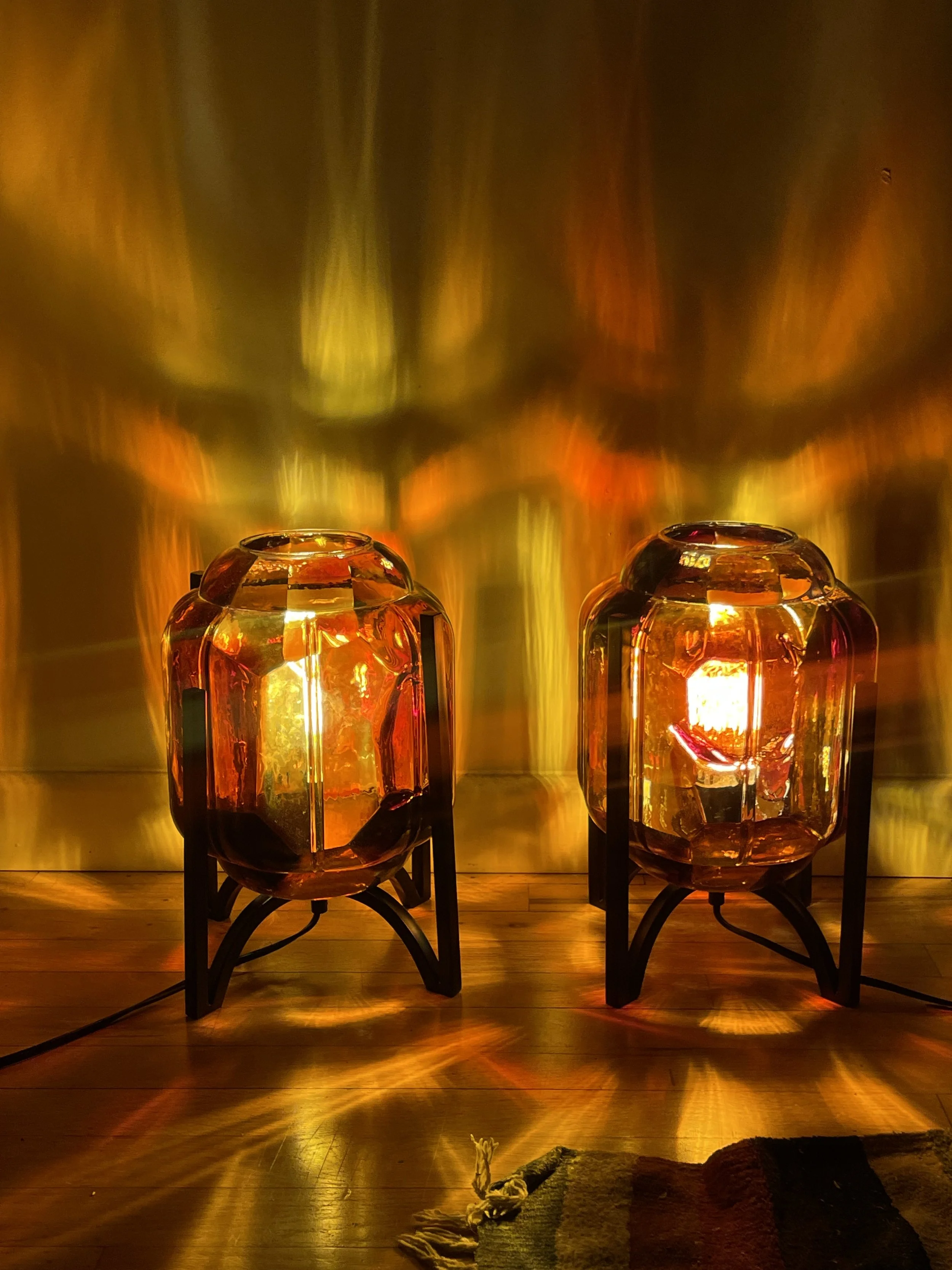 Two decorative glass and resin lamps with orange and yellow tones, casting colorful reflections and shadows on the wall and wooden floor.
