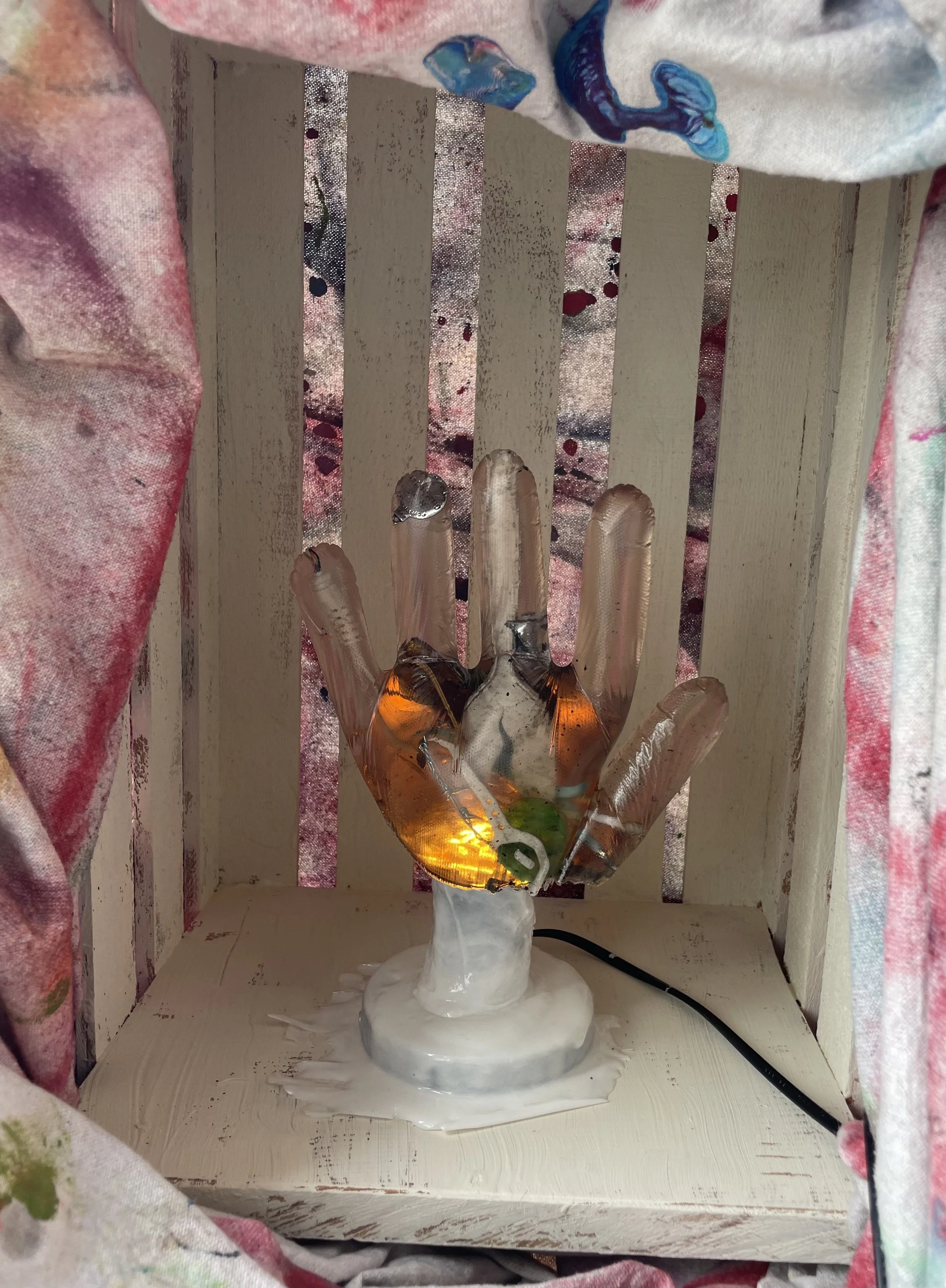 A resin light sculpture of a hand with trash inside, placed on a white wooden surface inside a small wooden frame, with fabric curtains surrounding it.