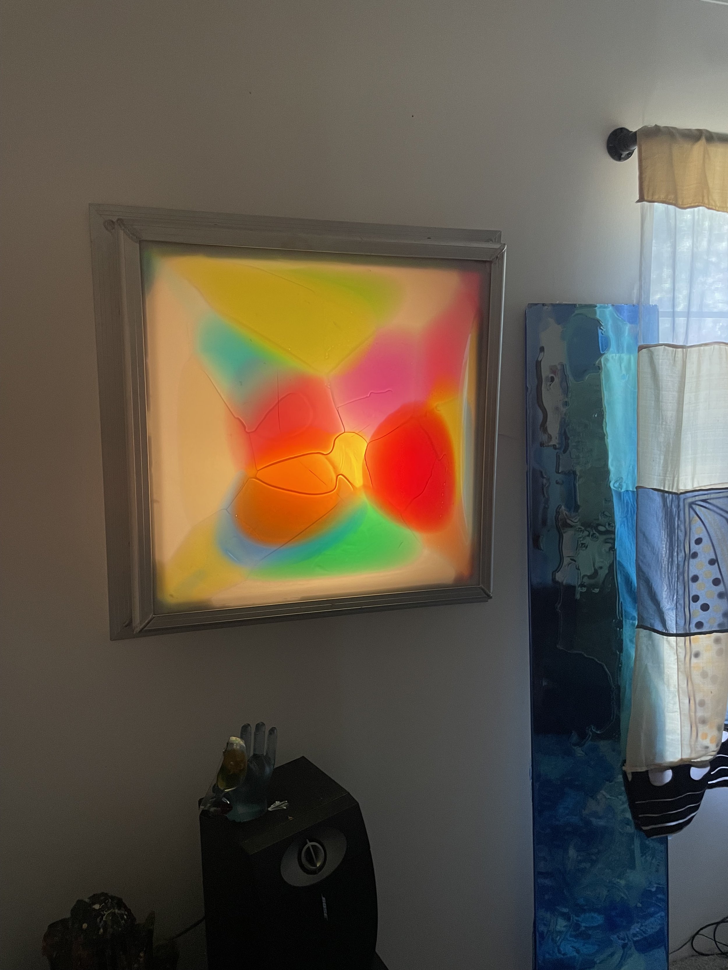 Colorful abstract resin light art with red, yellow, green, blue, and pink hues, framed on a wall next to blue and white curtains.