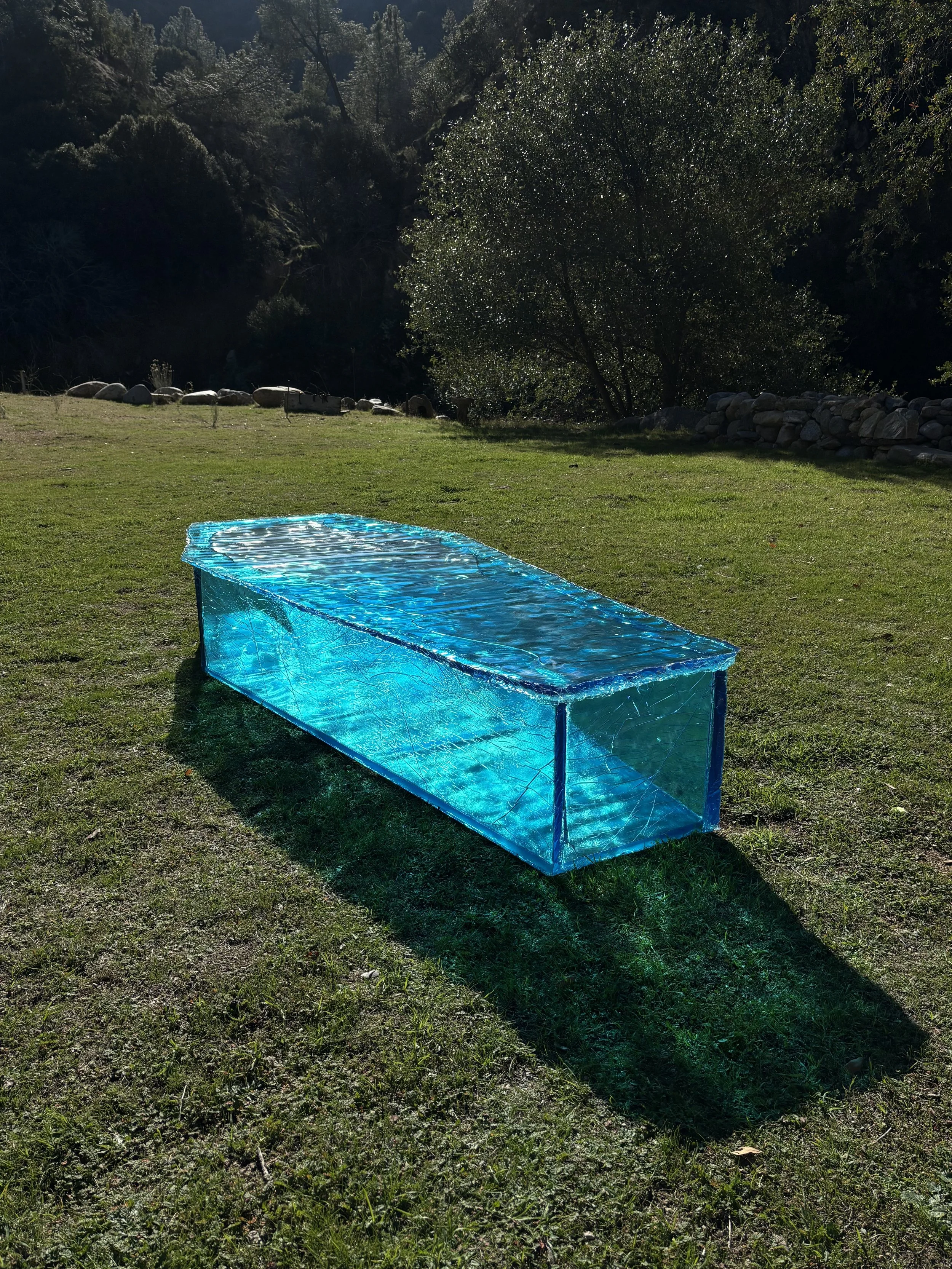 A clear blue glass-like resin coffin set up on a grassy lawn outdoors, with trees and rocks in the background.