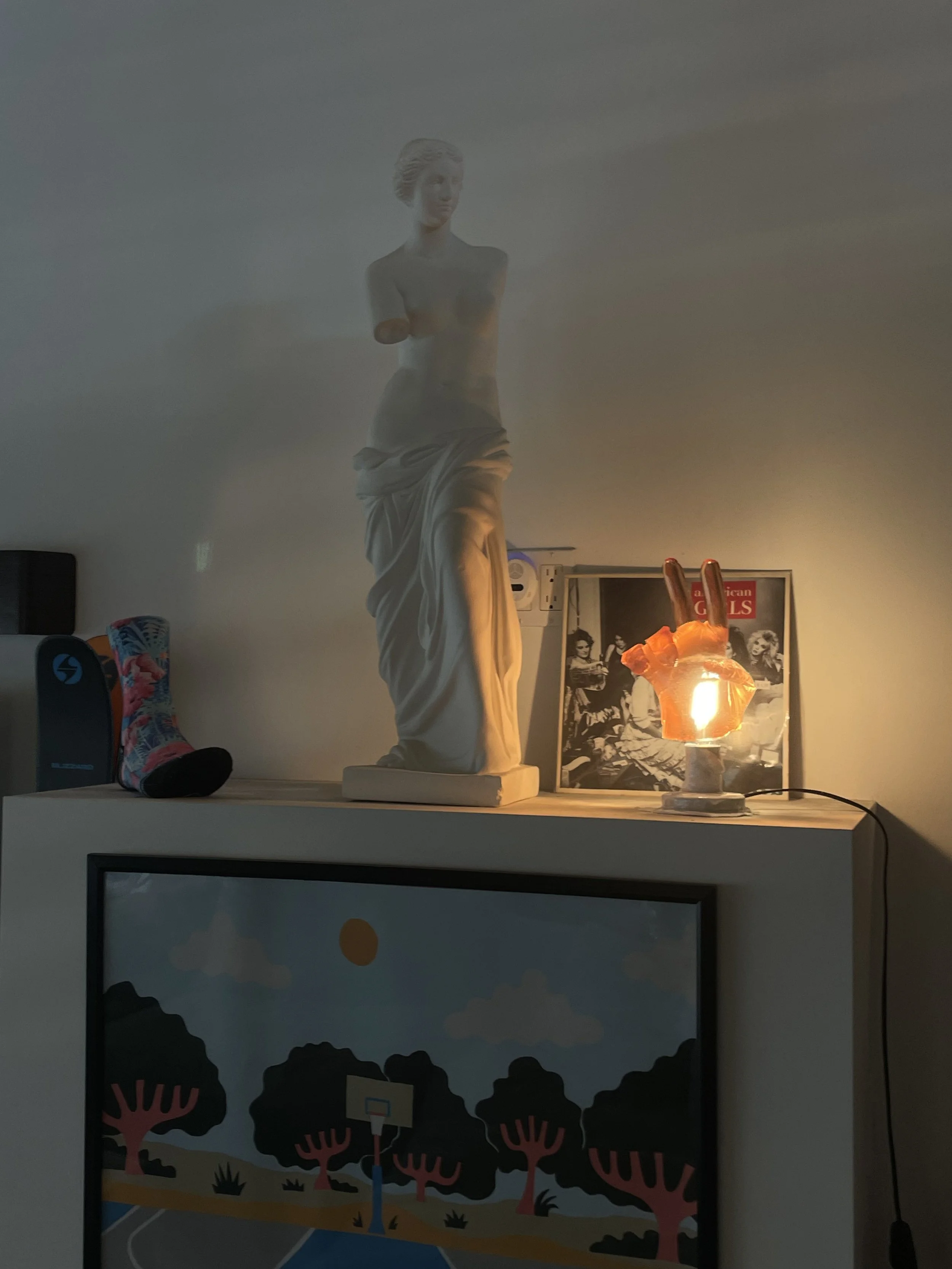 A shelf with a classical-style female sculpture, a peace sign shaped resin table lamp, a framed black-and-white photograph, and a sock on the left side. The lamp illuminates, casting a glow on the objects.
