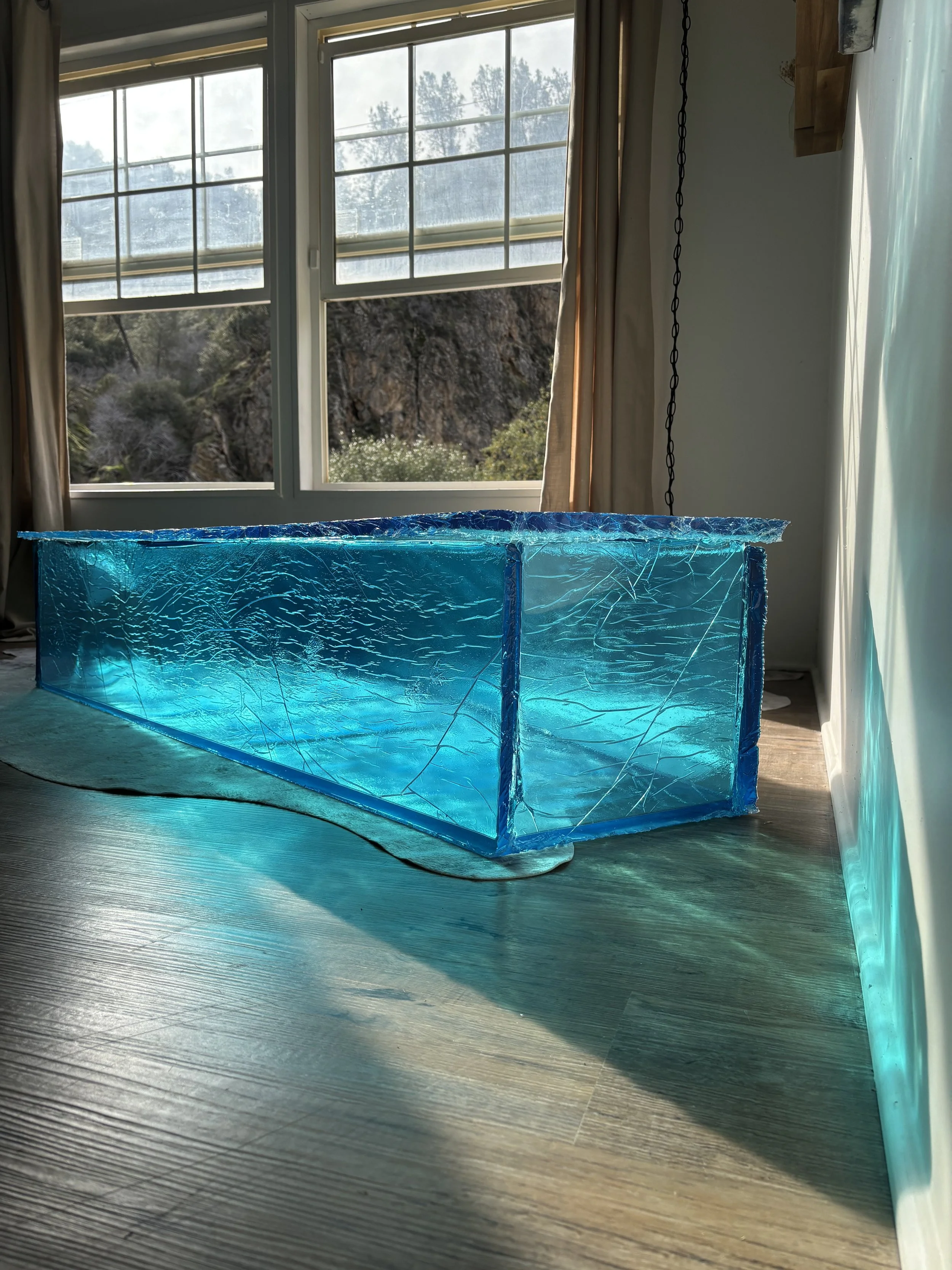 A blue ice-like resin coffin structure inside a room near large window with curtains, sunlight, and outdoor trees and mountains visible.