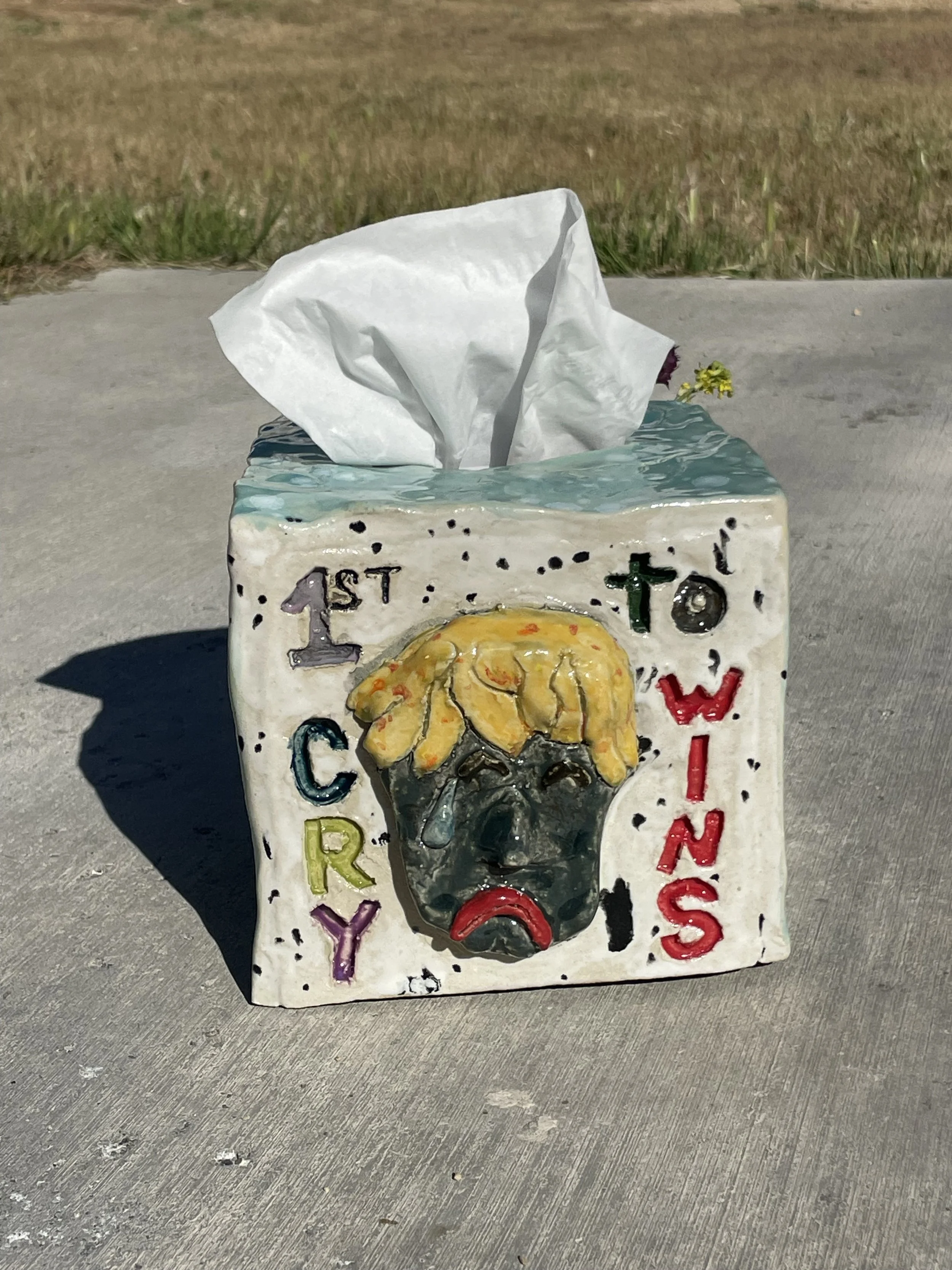 Decorative tissue box cover with a sad face and stylized blonde hair, surrounded by the words '1st Cry to WINS' in colorful letters, placed on concrete outdoors.