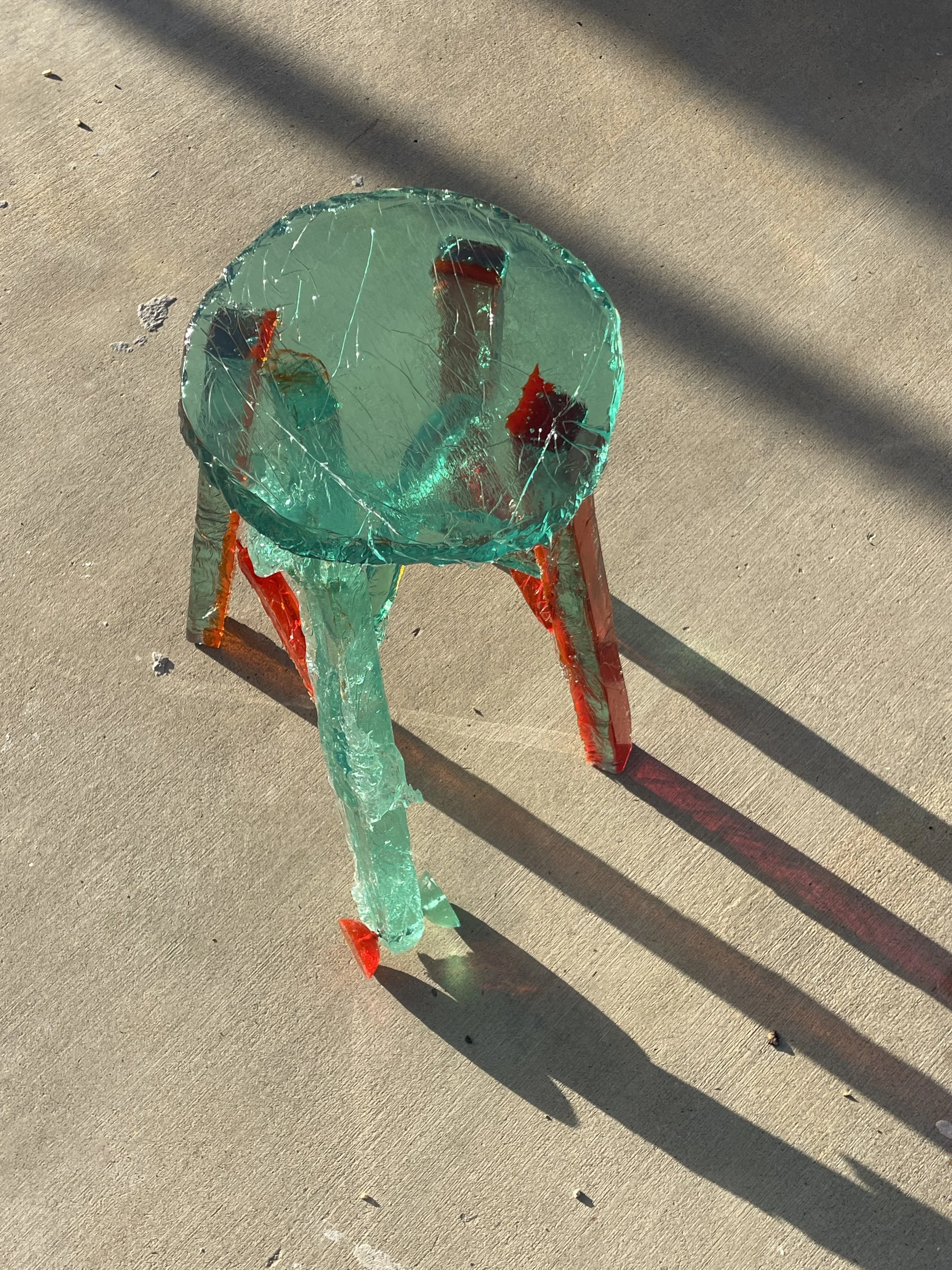 Broken glass sculpture resembling a small table with red and green accents, casting a shadow on concrete ground.