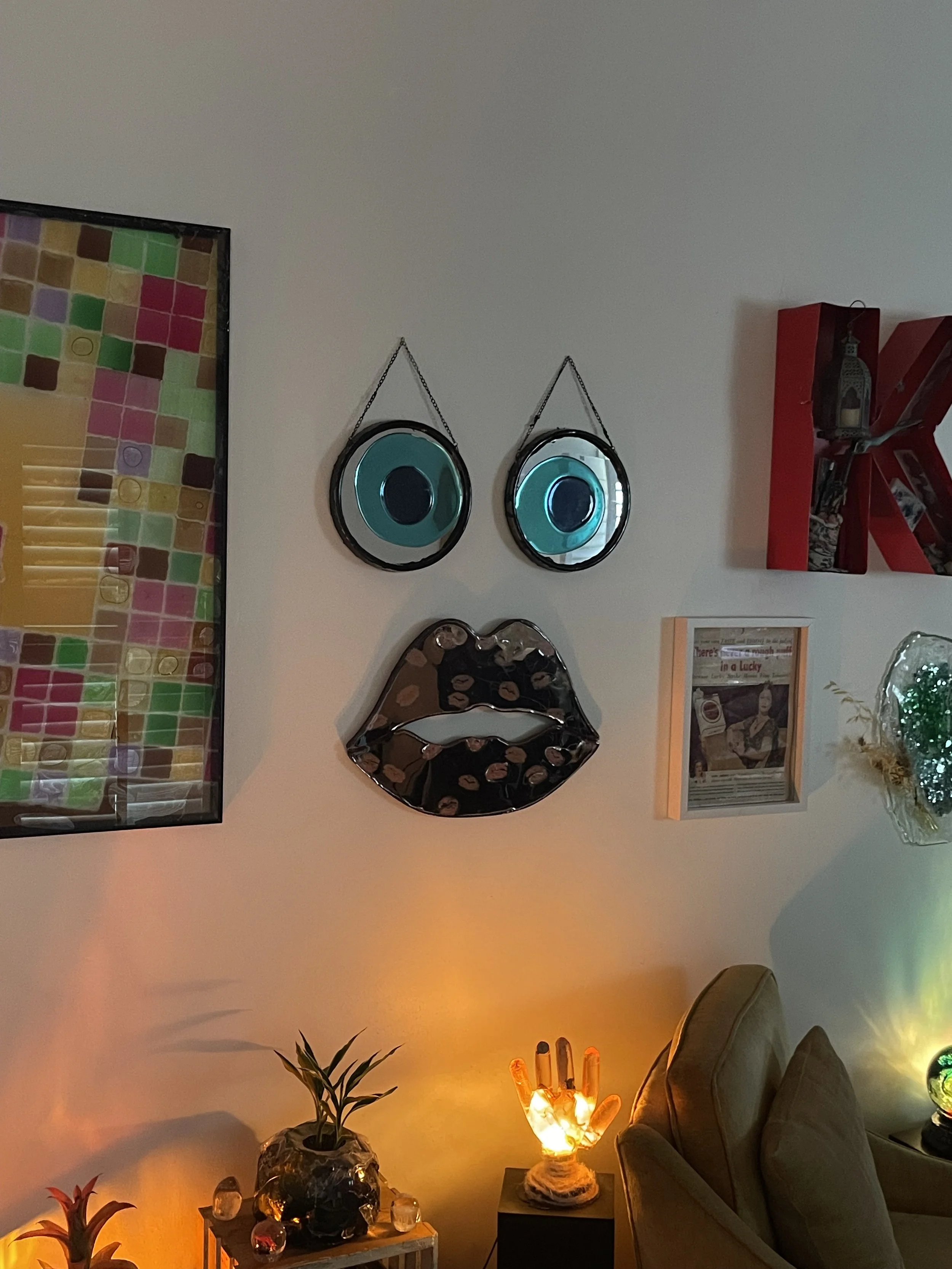 Decorative wall art featuring a face with two large blue eyes and glossy black lips with kiss marks, surrounded by framed pictures and plants in a cozy living room.