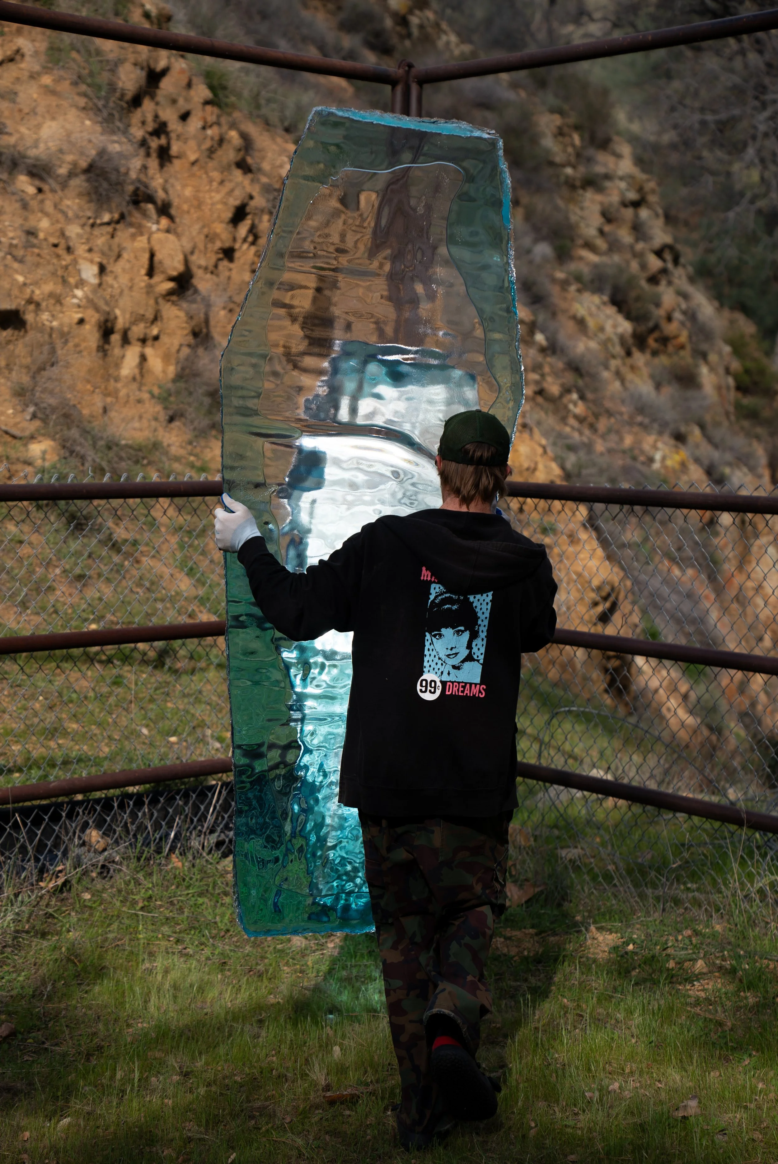 Person with a dark hoodie, camouflage pants, and a green cap holding a large, resin coffin lid, standing near a chain-link fence with a rocky hillside in the background.