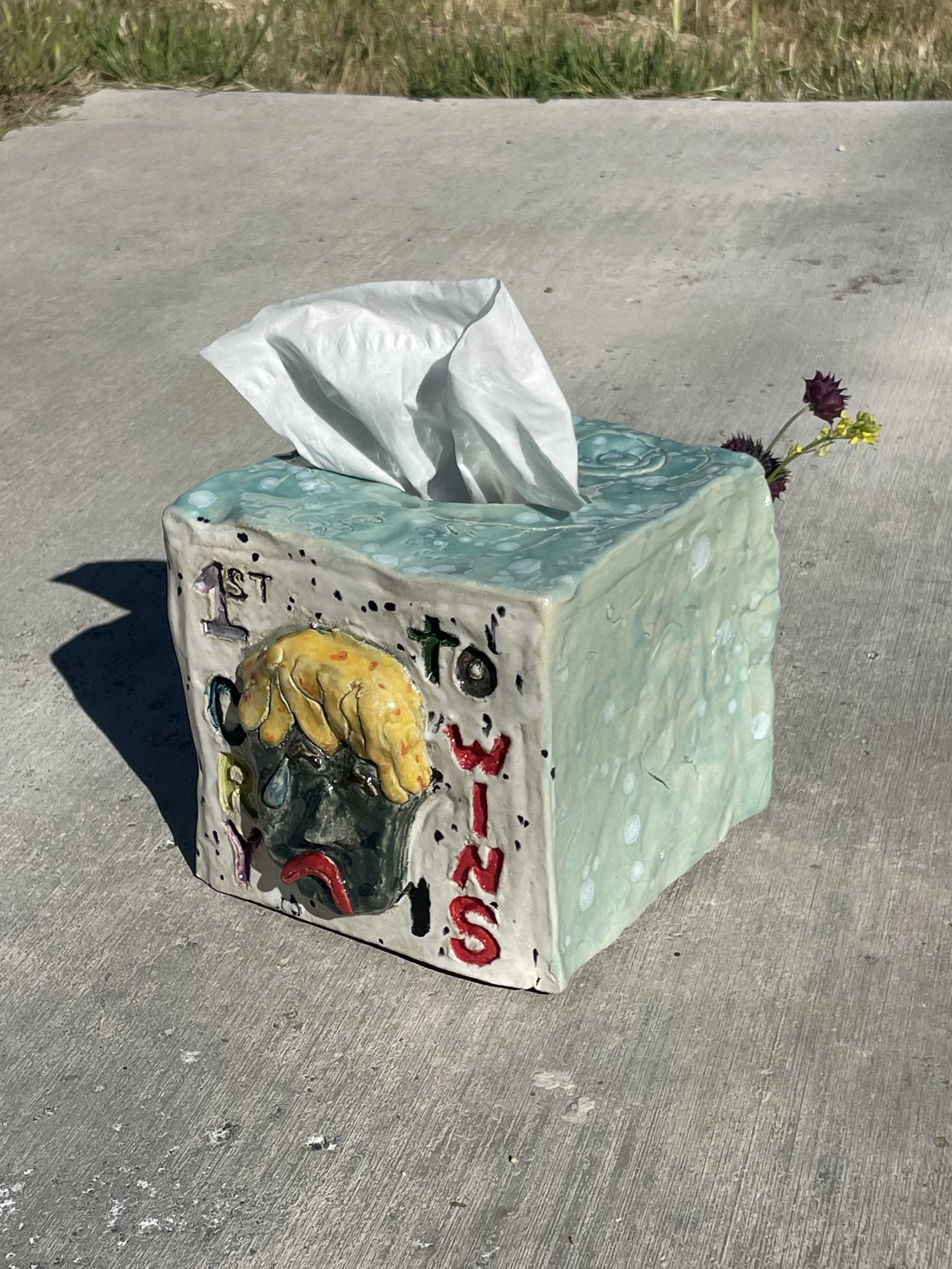 A ceramic tissue box cover decorated with a colorful abstract face and the word 'LUSO' on one side, placed on a concrete surface with some flowers peeking out of the side.