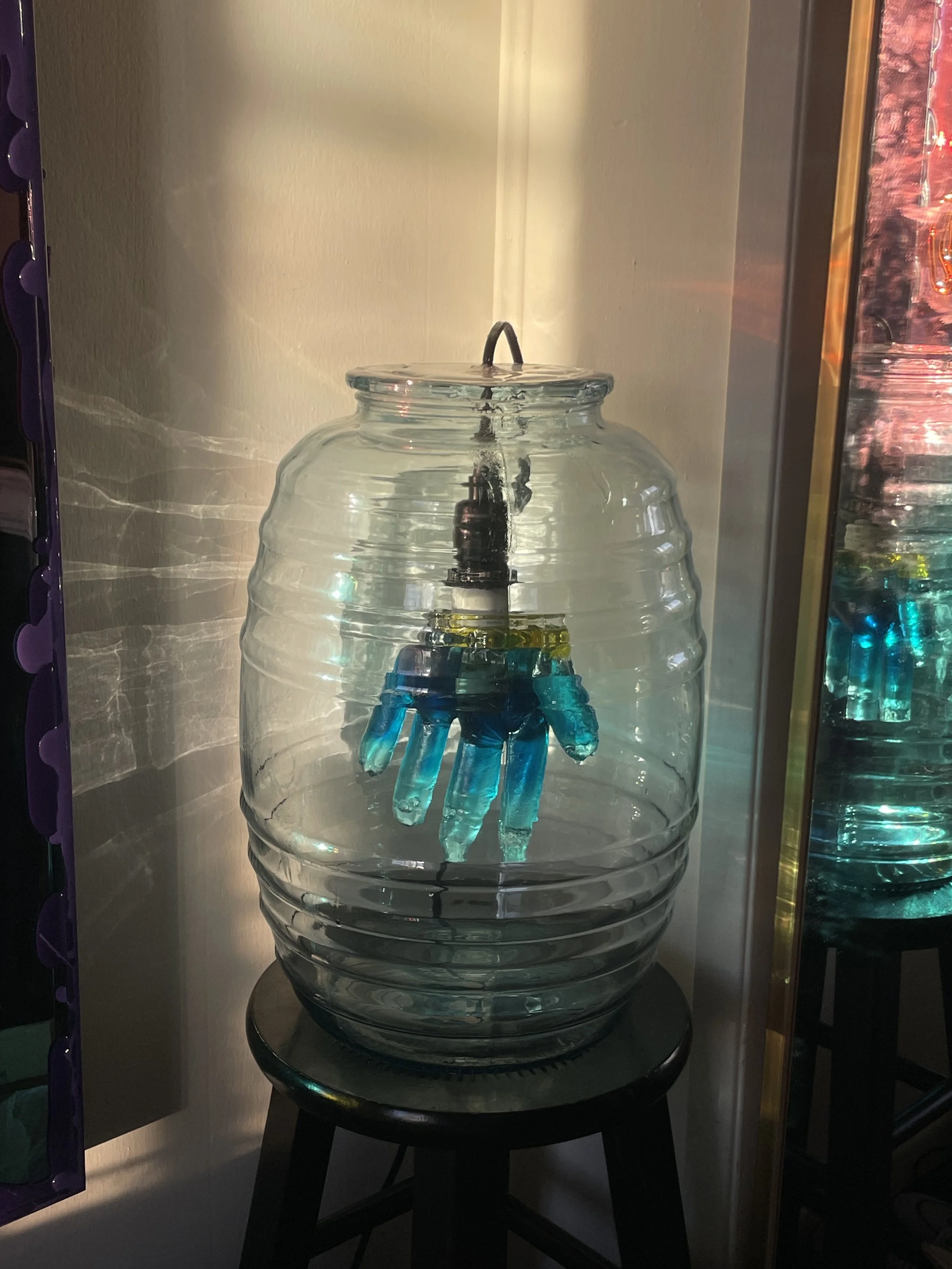 A glass jar with a human-hand-shaped resin sculpture inside, connected to a black cord.