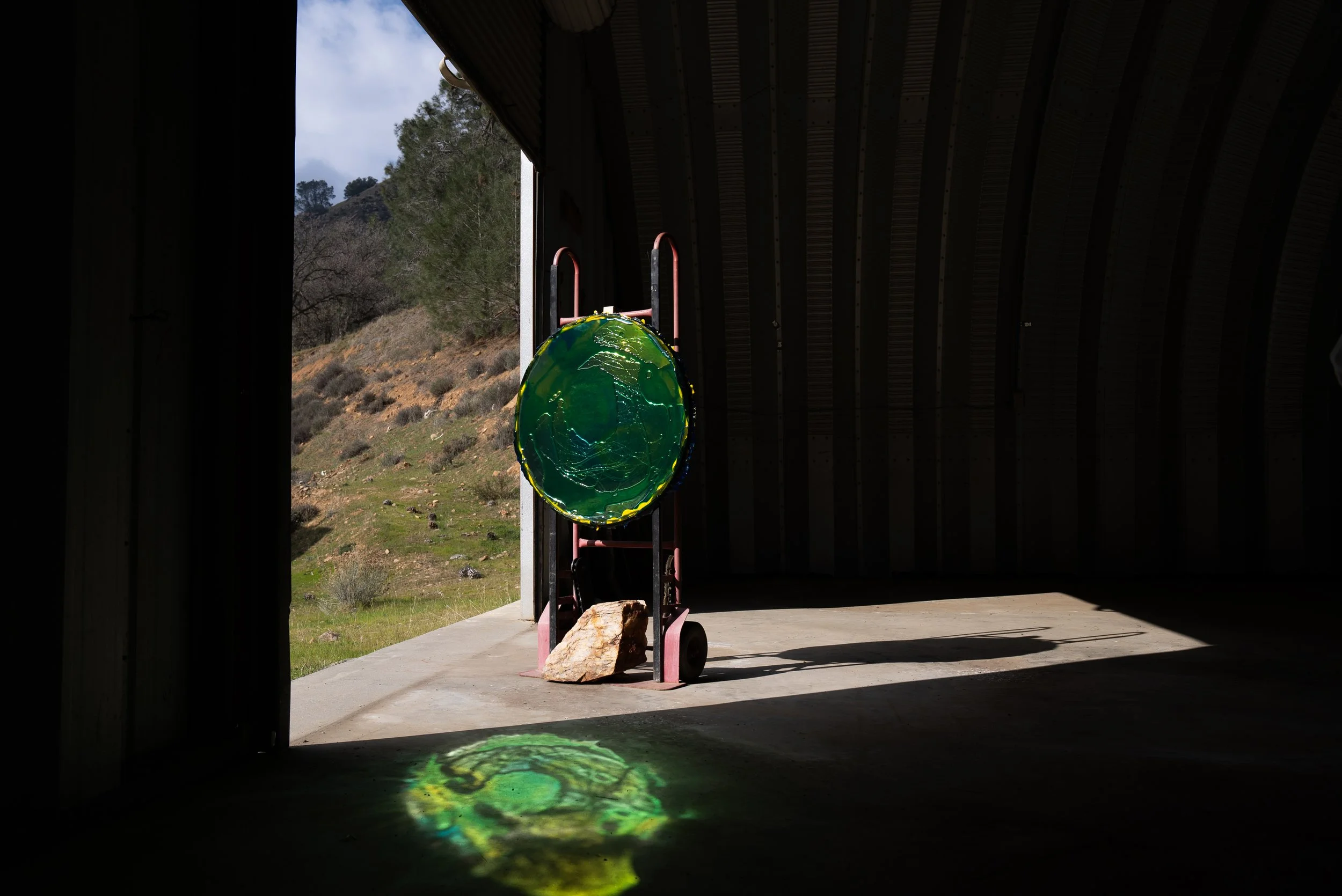 Looking out of a barn or shed doorway onto a hillside with trees and grass, with a green resin mirror hanging on a dolly inside, casting a reflection on the floor.