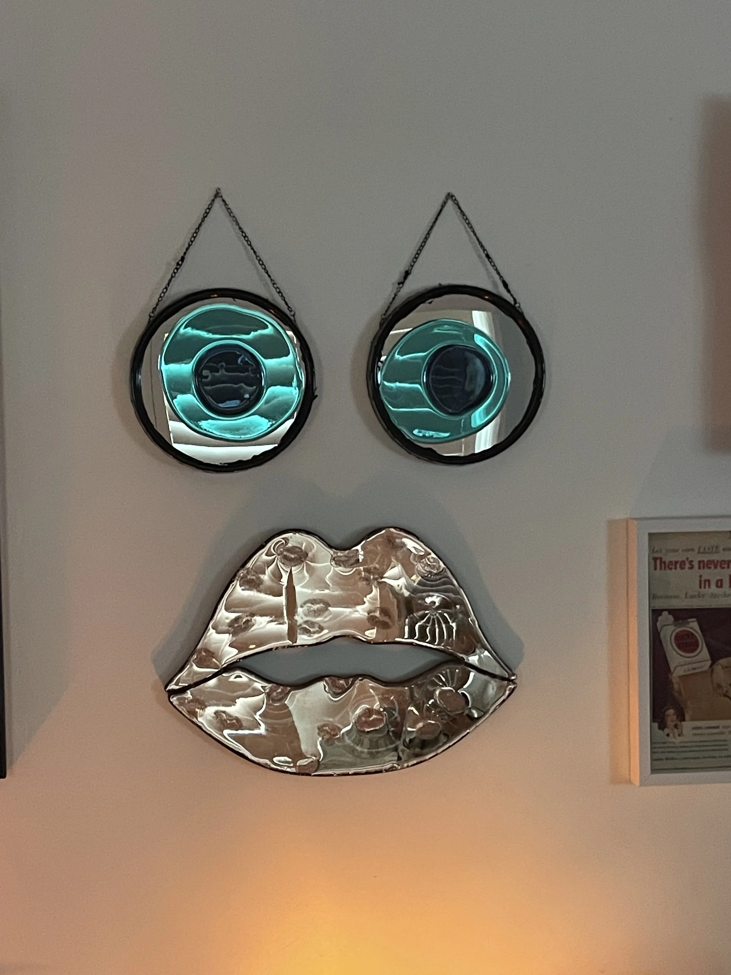 Decorative wall art featuring two round mirrors with neon blue resin as eyes and a large metallic lips sculpture beneath, all hanging on a plain wall.