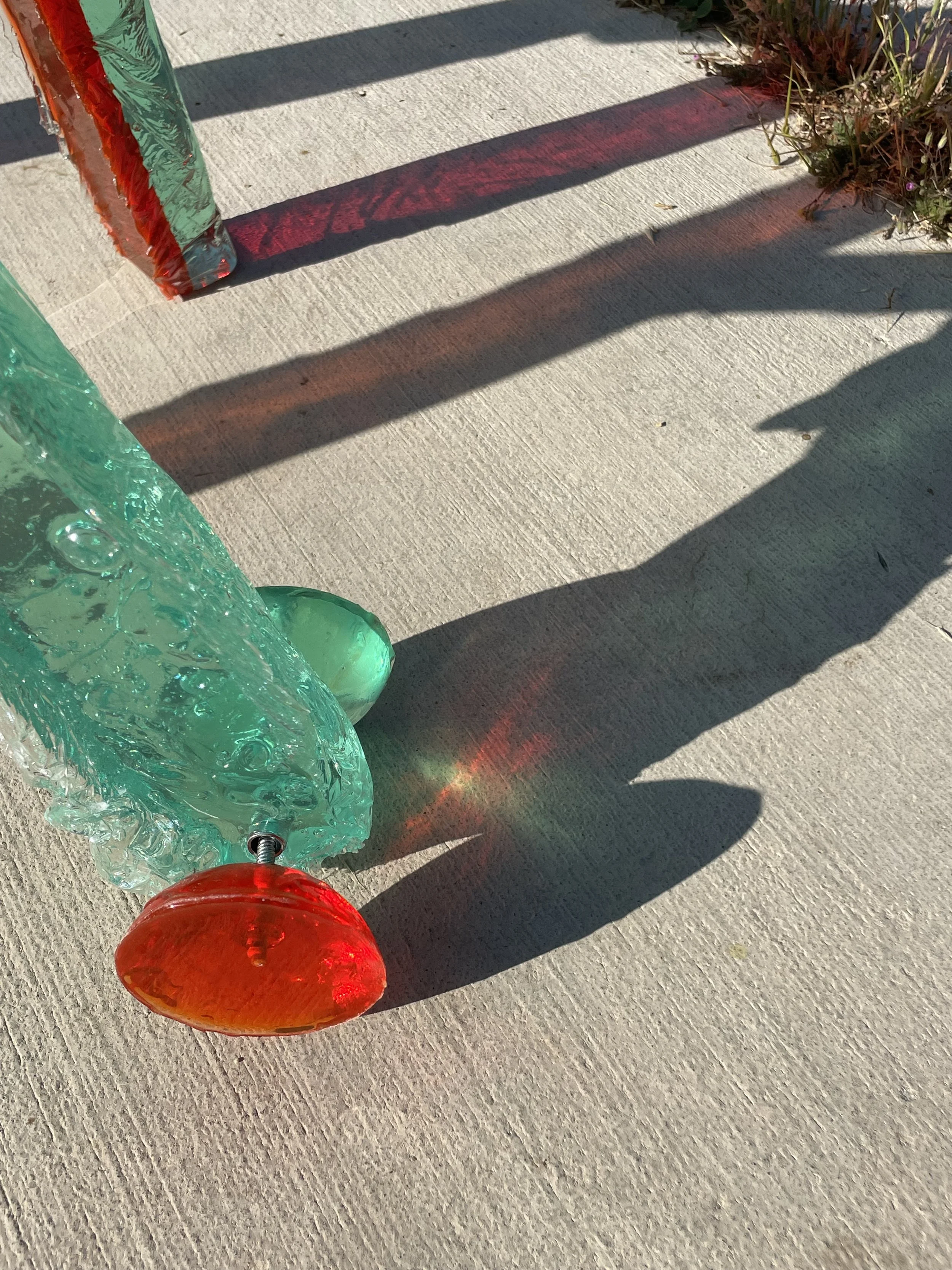 Glass sculpture with red, green, and clear elements casting colorful shadows on concrete ground, with small plants nearby.