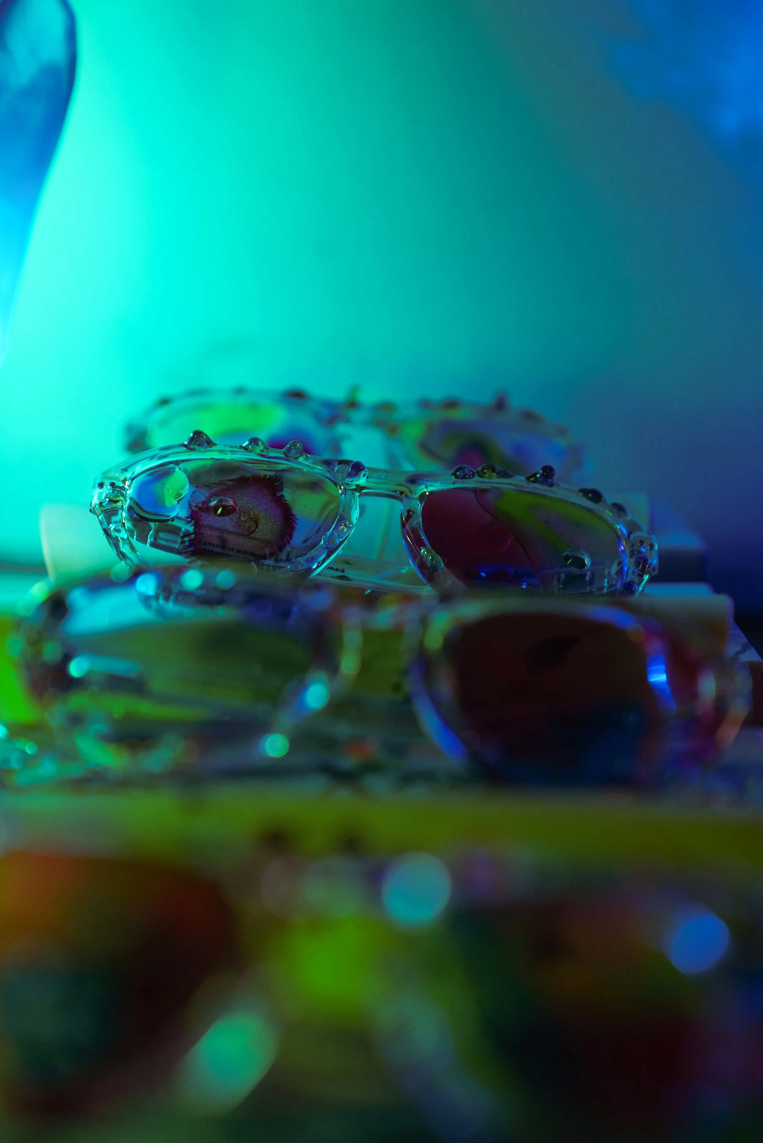 Close-up of colorful sunglasses with resin droplets on the frames, illuminated by vibrant blue, green, and purple lighting.