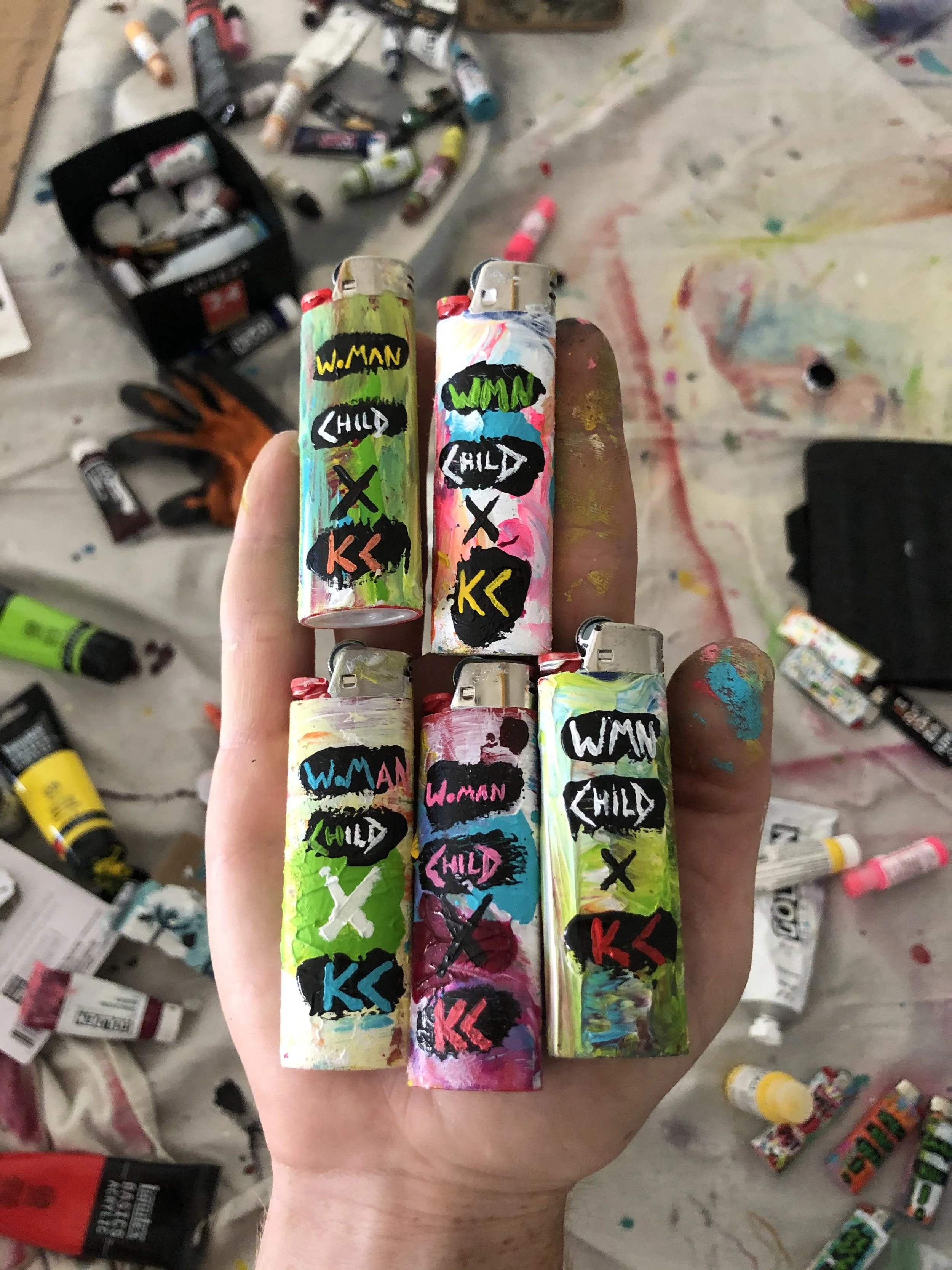 Six painted metal lighters with colorful abstract designs and labels reading 'Woman' and 'Child' with X marks between them, held in a person's hand against a painter's workspace background with paint tubes and brushes.