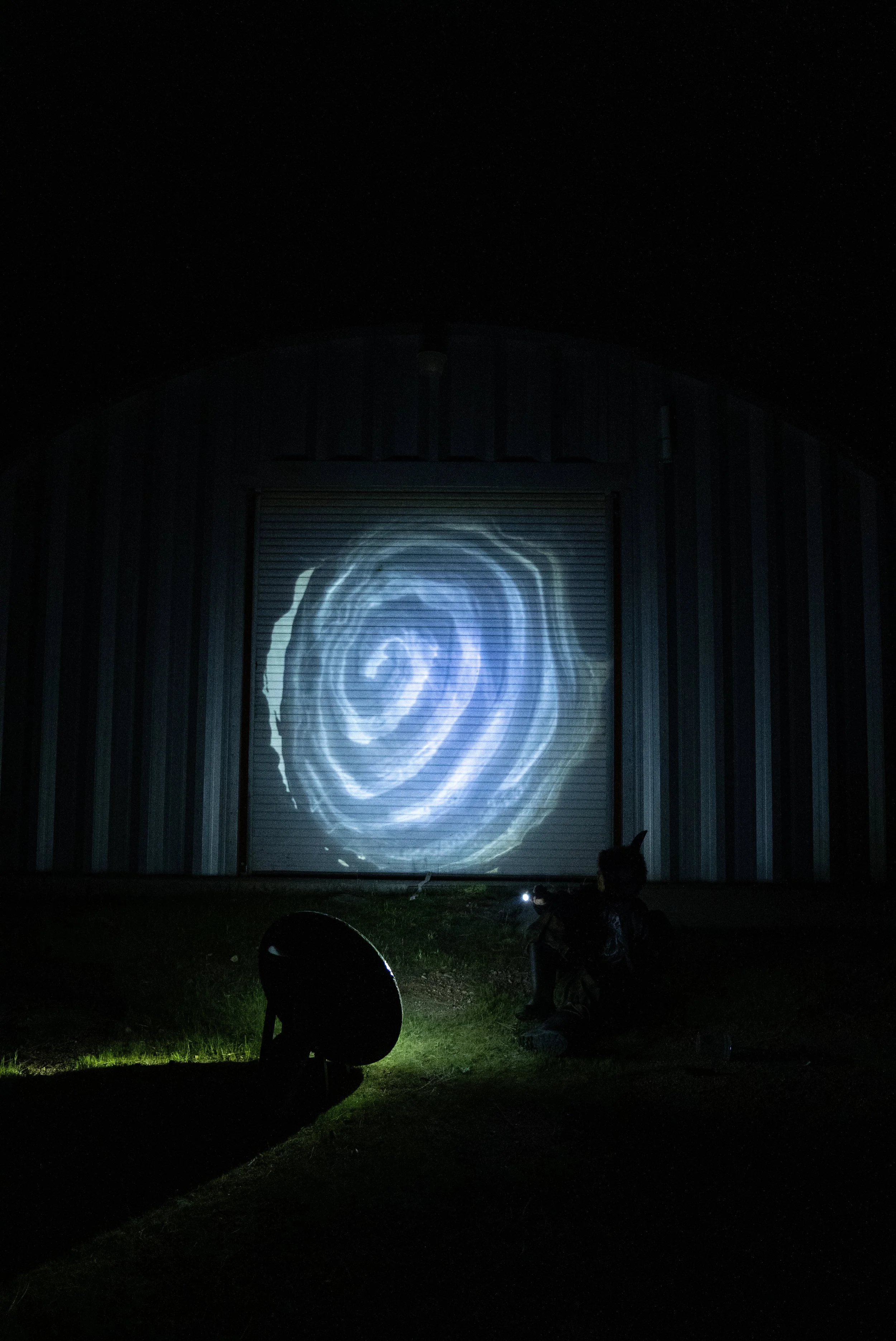 A dark outdoor scene shows a person sitting on the ground pointing a flashlight at a circular resin mirror, bouncing the reflections onto a barn door with a swirling light projection resembling an eye or spiral galaxy.