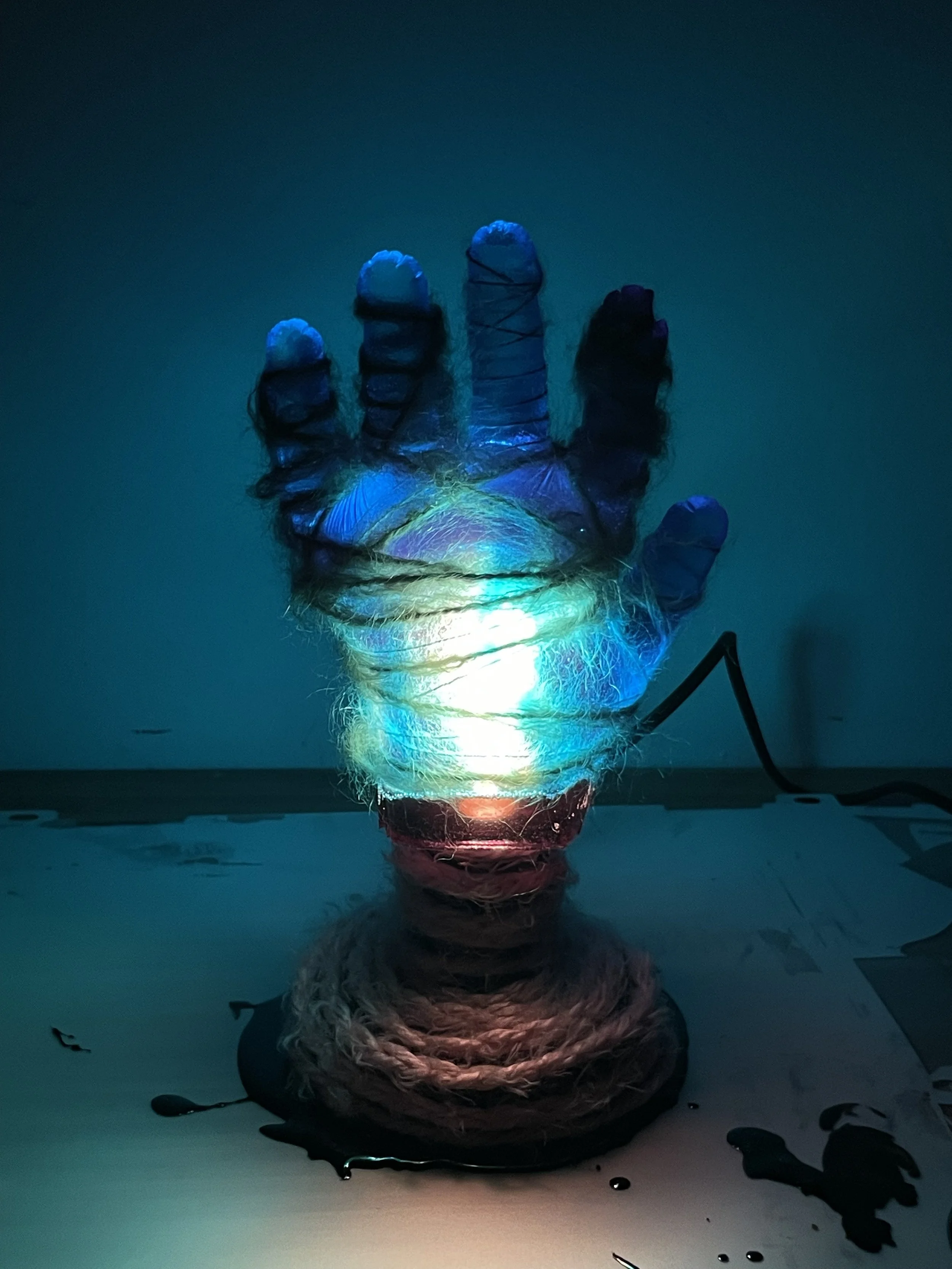 A colorful illuminated resin sculpture of a human hand made from wrapped string or yarn, placed on a dark surface with some spilled black paint or ink