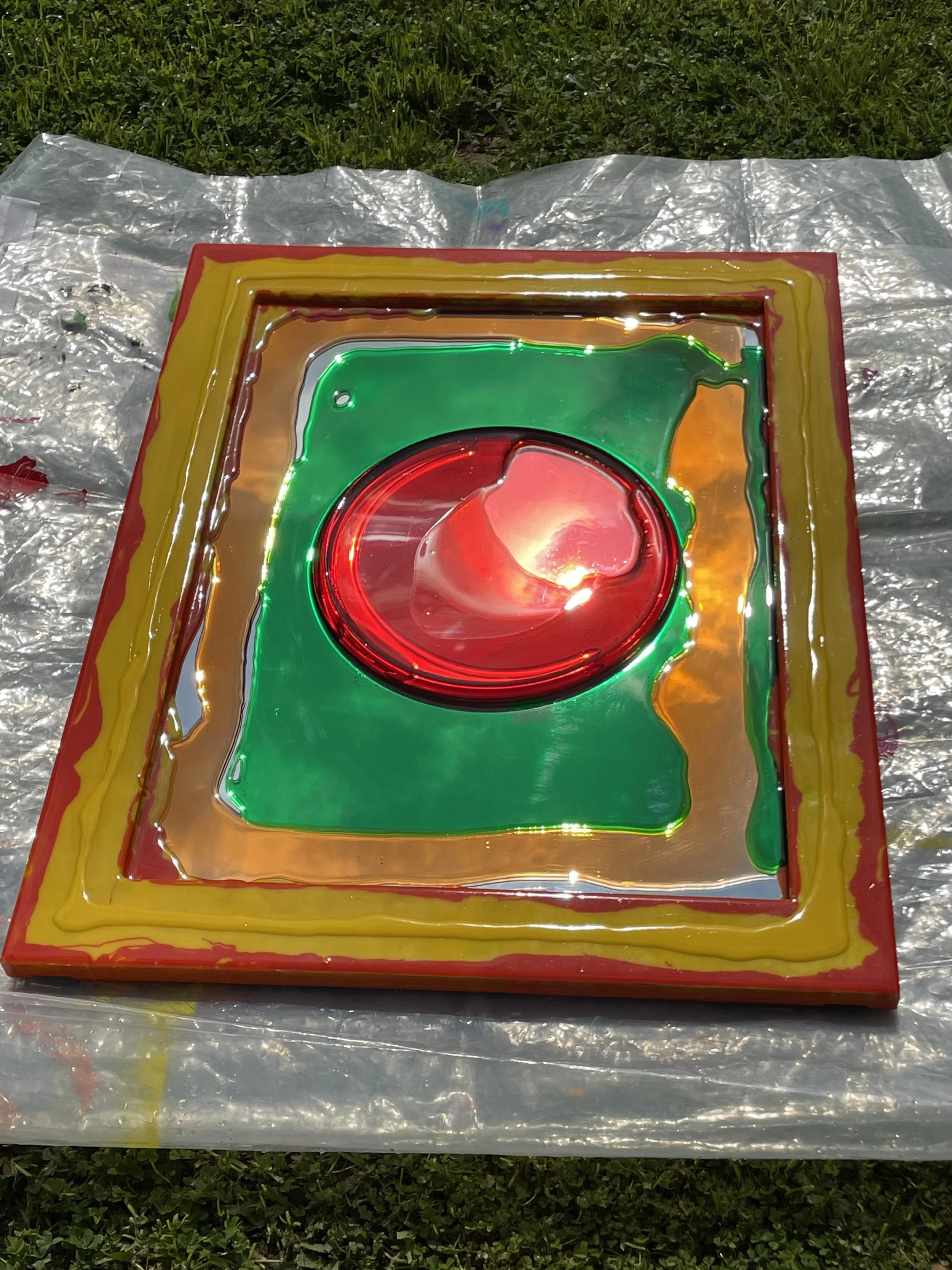 Colorful resin mirror with red, green, yellow, and orange resin, lying on plastic on grass outside.