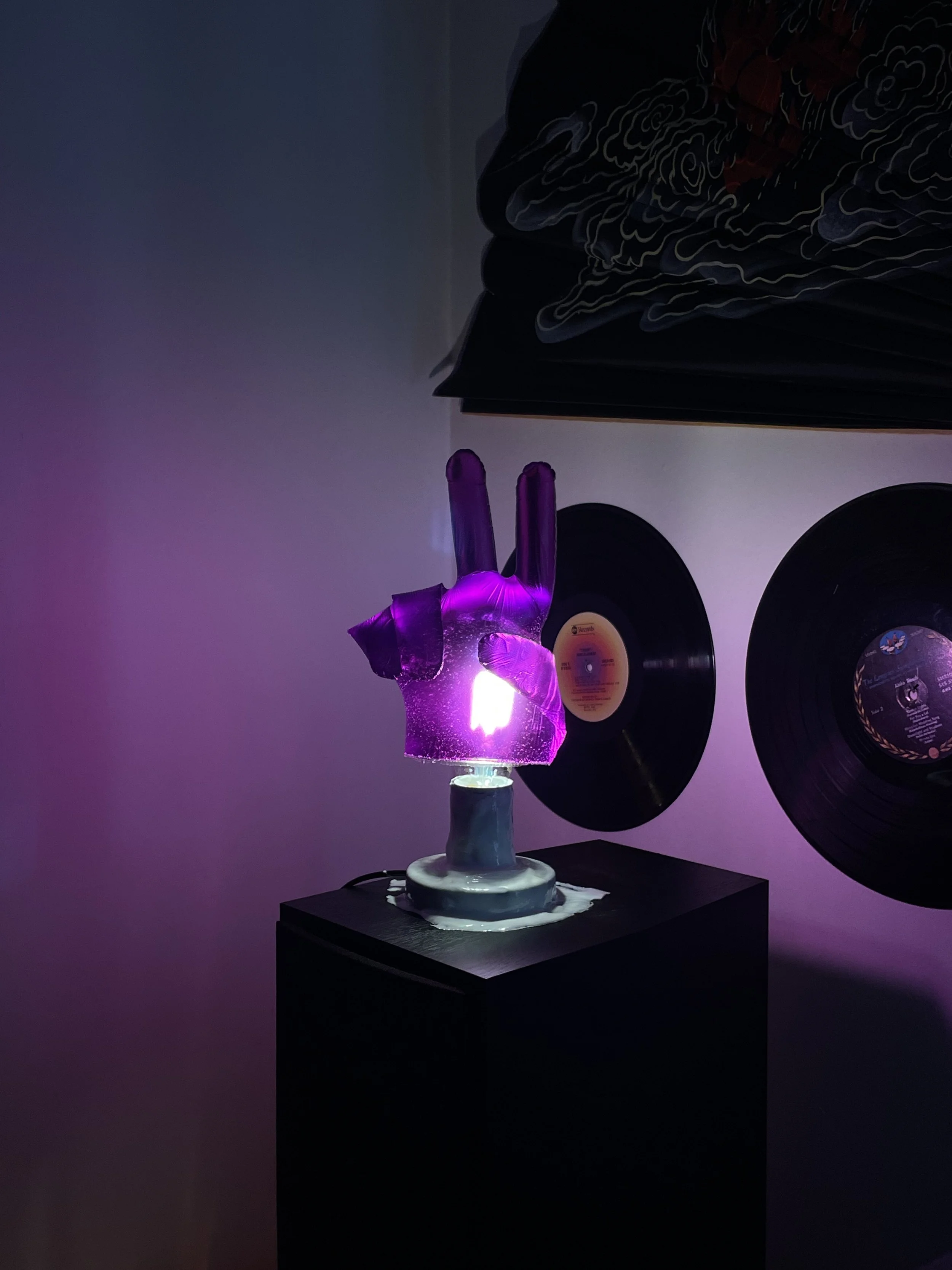 A quirky resin lamp in the shape of a purple hand making a peace sign, illuminated with purple light, on a black pedestal with two vinyl records hanging on the wall behind it.