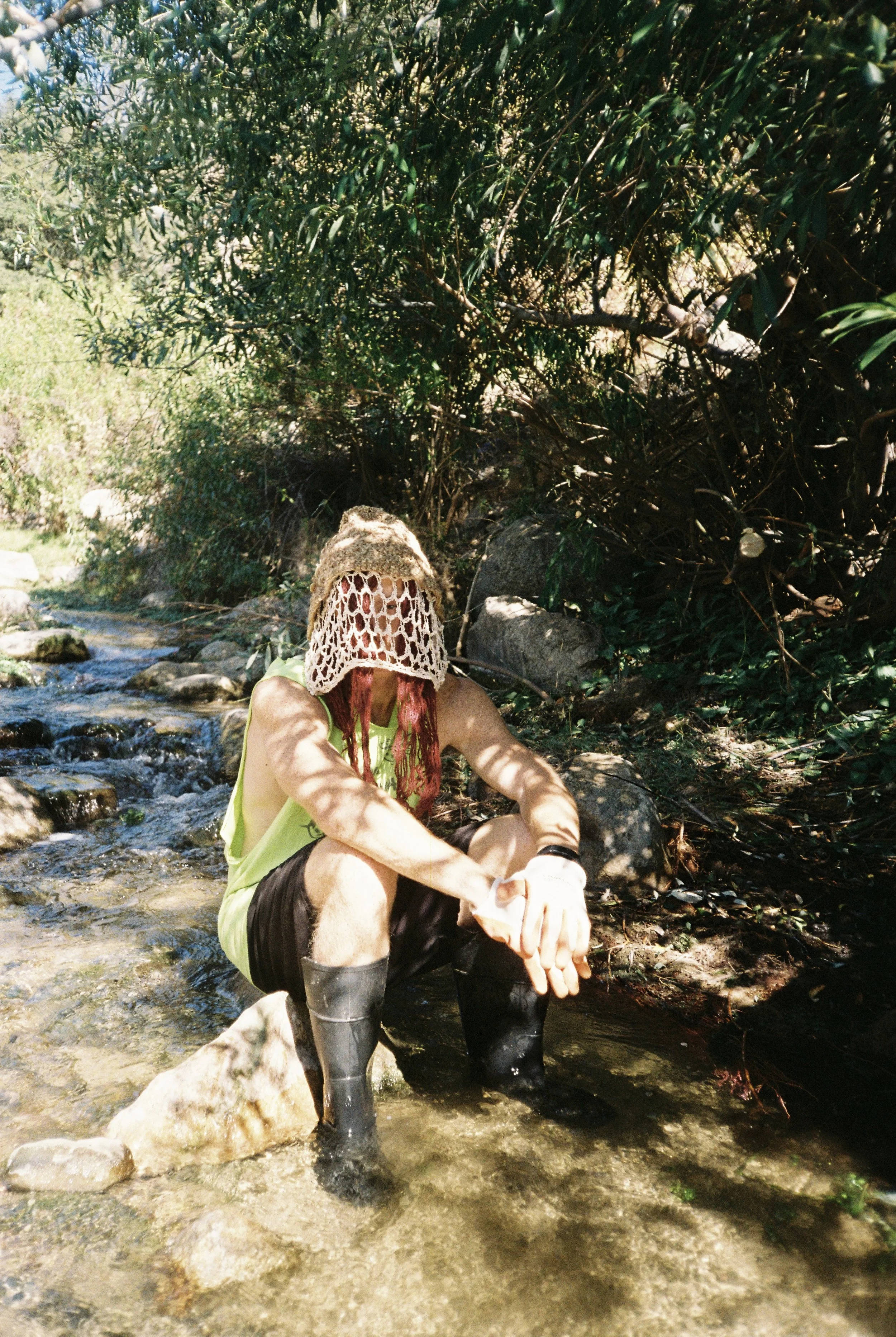 Kendall Cleveland sitting in a shallow stream, wearing an Auntie Flew crocheted mask, neon green shirt, black shorts, and tall rubber boots, surrounded by greenery and rocks.