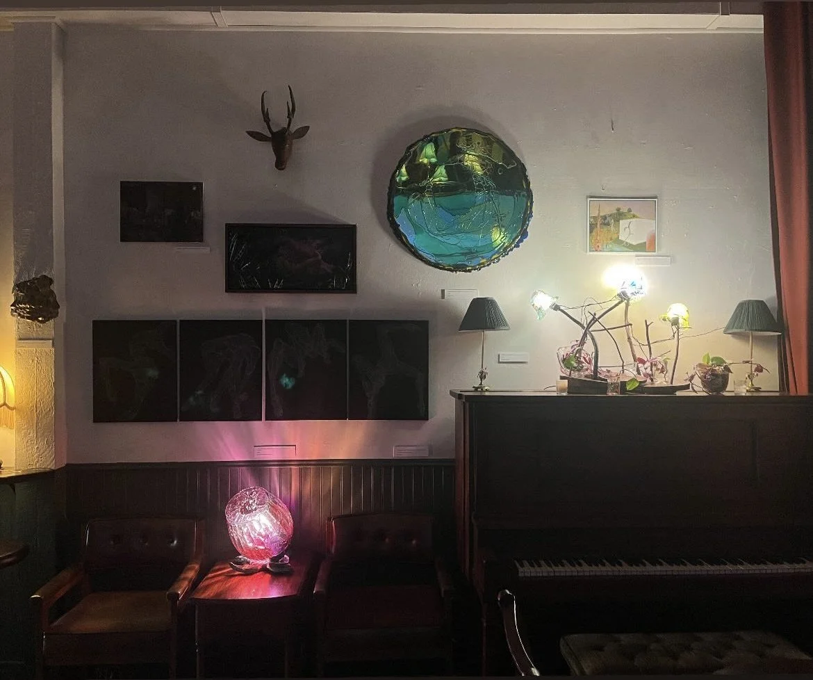 Interior of a cozy room with a black upright piano, a small table with a pink illuminated decorative lamp, two dark brown chairs, a wall-mounted deer head, various framed artworks, a colorful glass wall art, and a string of bright lights on top of th