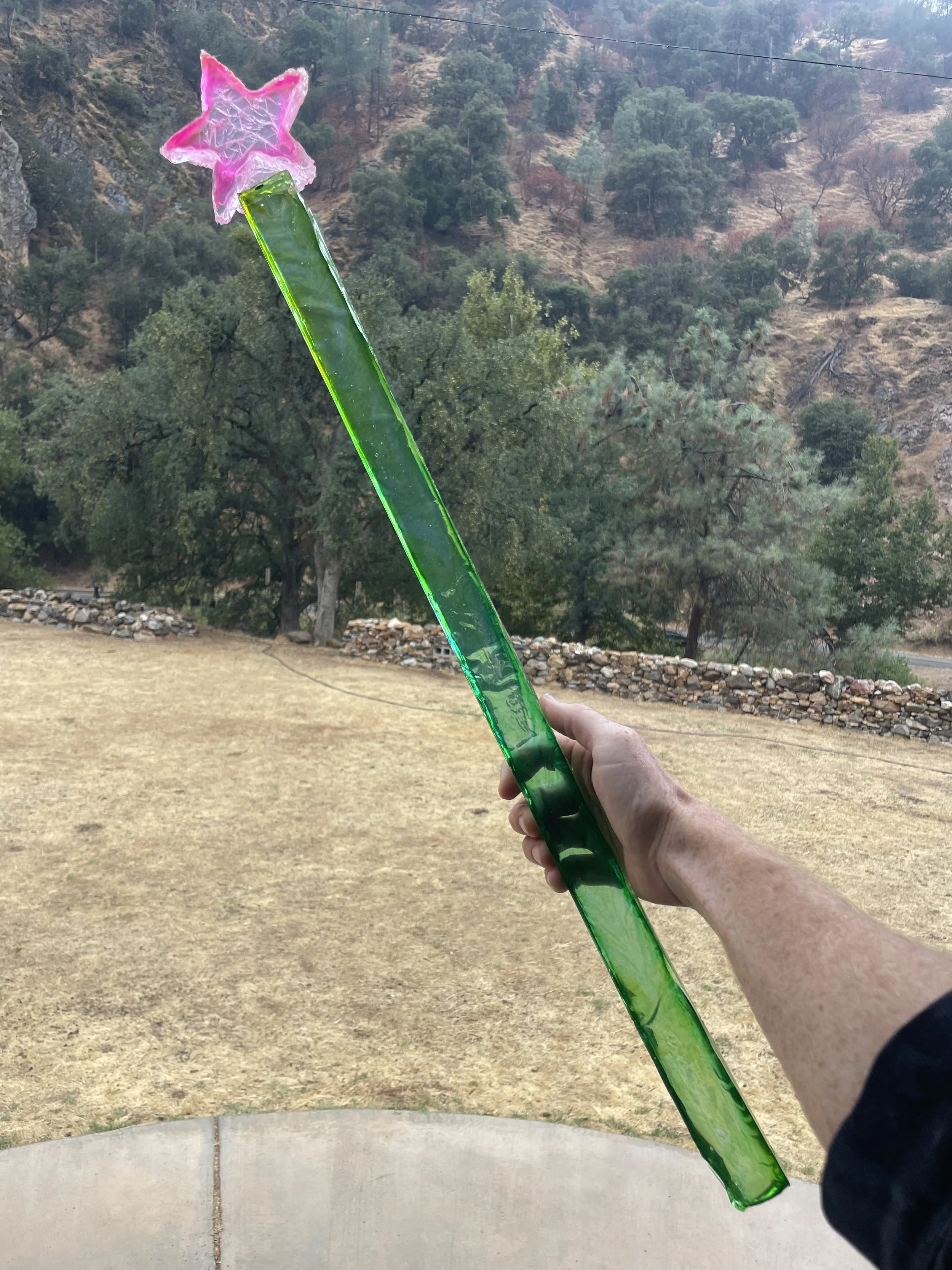Hand holding a green magical resin wand with a pink star at the tip against an outdoor background of trees and hills.