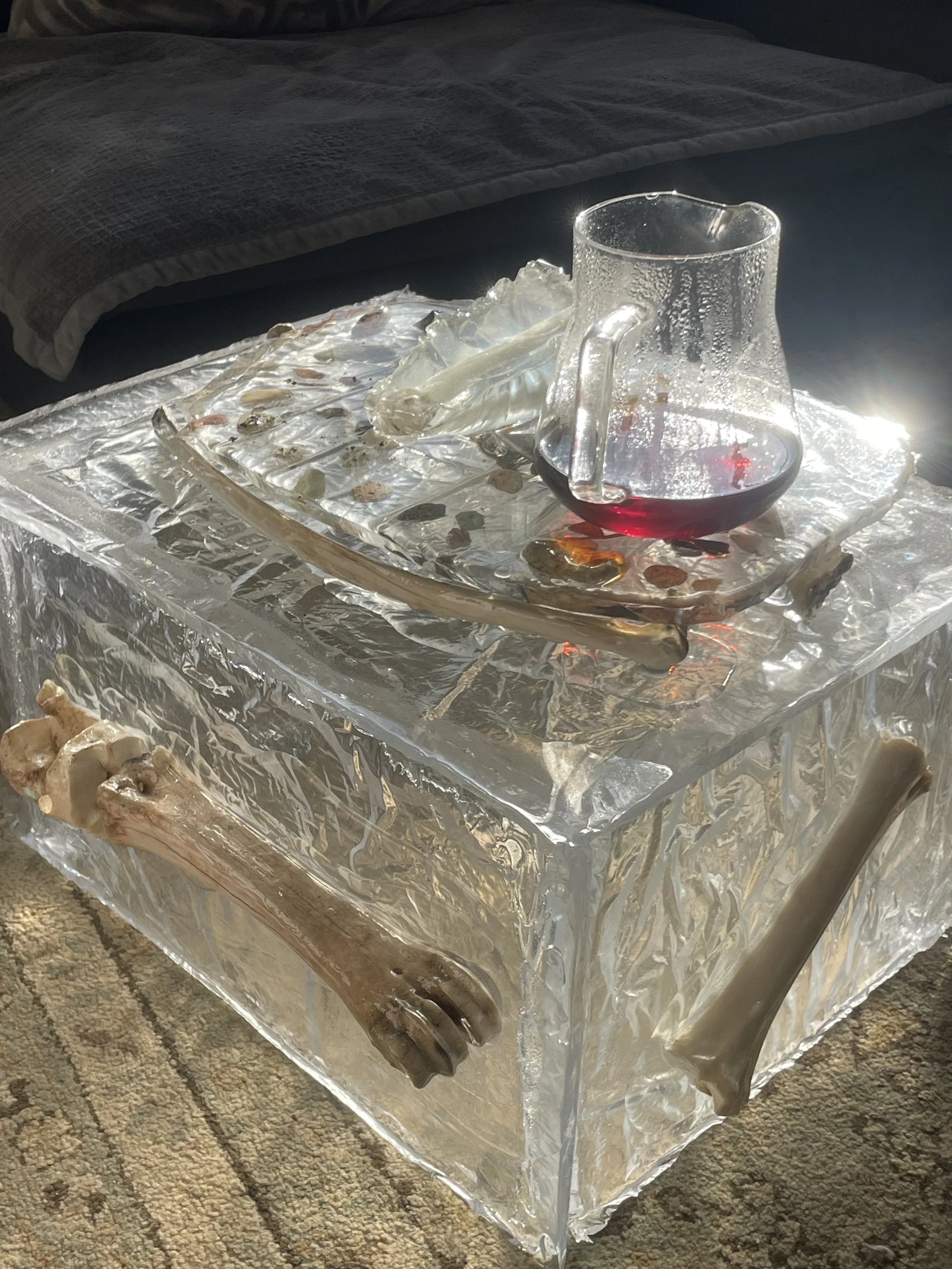 Glass pitcher with red liquid, placed on a transparent resin box with bones and stones around it in a room with a bed and carpet.