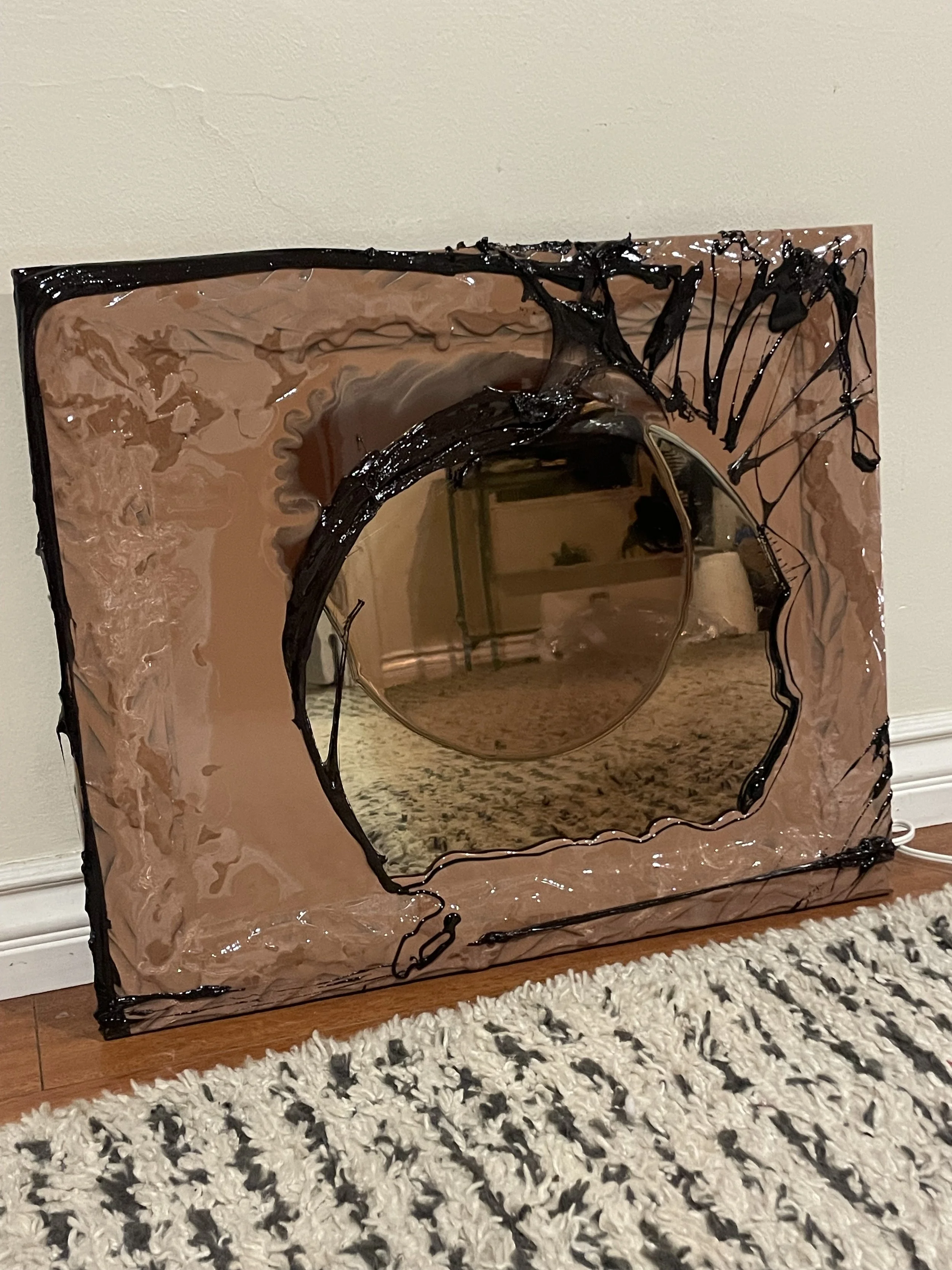 A decorative resin mirror with a black and beige frame leaning against a wall on a wooden floor, reflecting a room with furniture and a patterned rug.