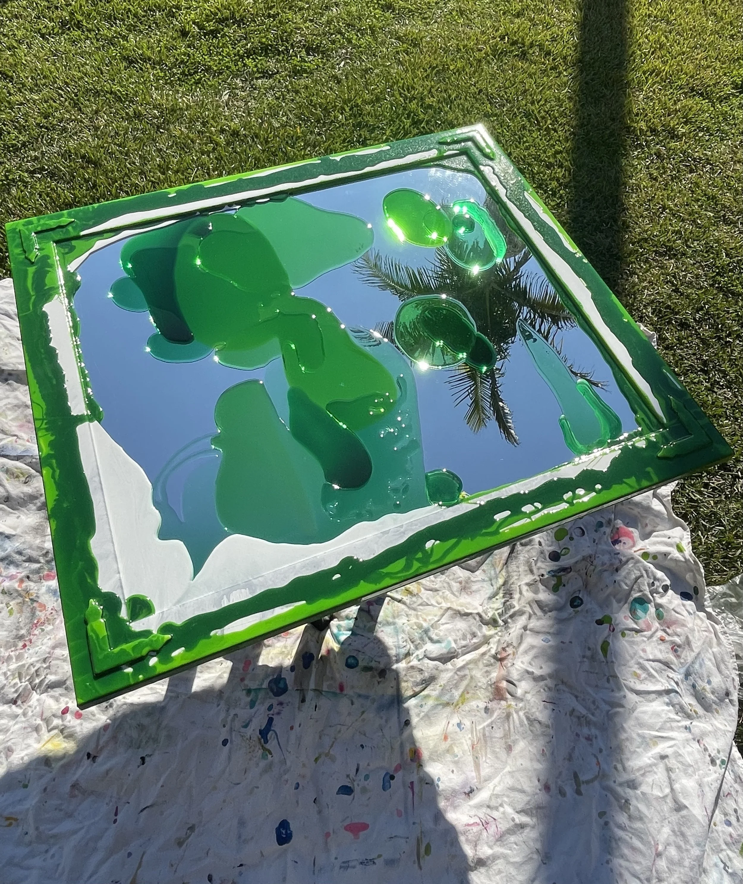A resin mirror with green and white colors, reflecting the sky and a palm tree in the reflection. The panel is outdoors on a white cloth on green grass.
